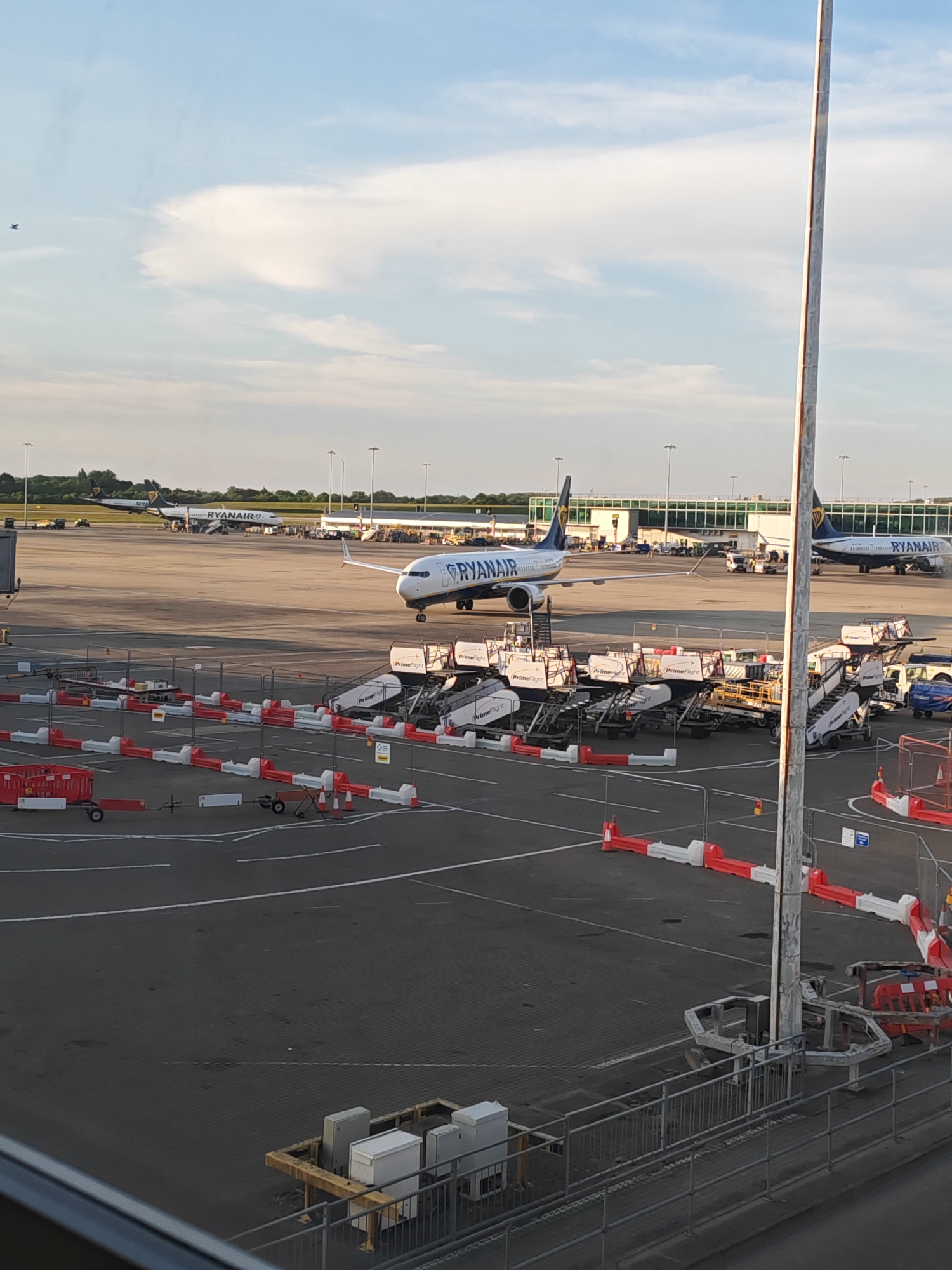 The image depicts an airport scene in Uttlesford, United Kingdom. Several Ryanair airplanes are visible on the tarmac, some appearing to be taxiing or stationary near terminal buildings. Numerous passenger boarding stairs, marked with "PrimeFlight," are situated in the foreground, along with various pieces of airport equipment. Red and white traffic barriers delineate areas within the airport's operational space. In the background, low-lying trees and other airport buildings can be seen. The sky is partly cloudy, suggesting daylight conditions.