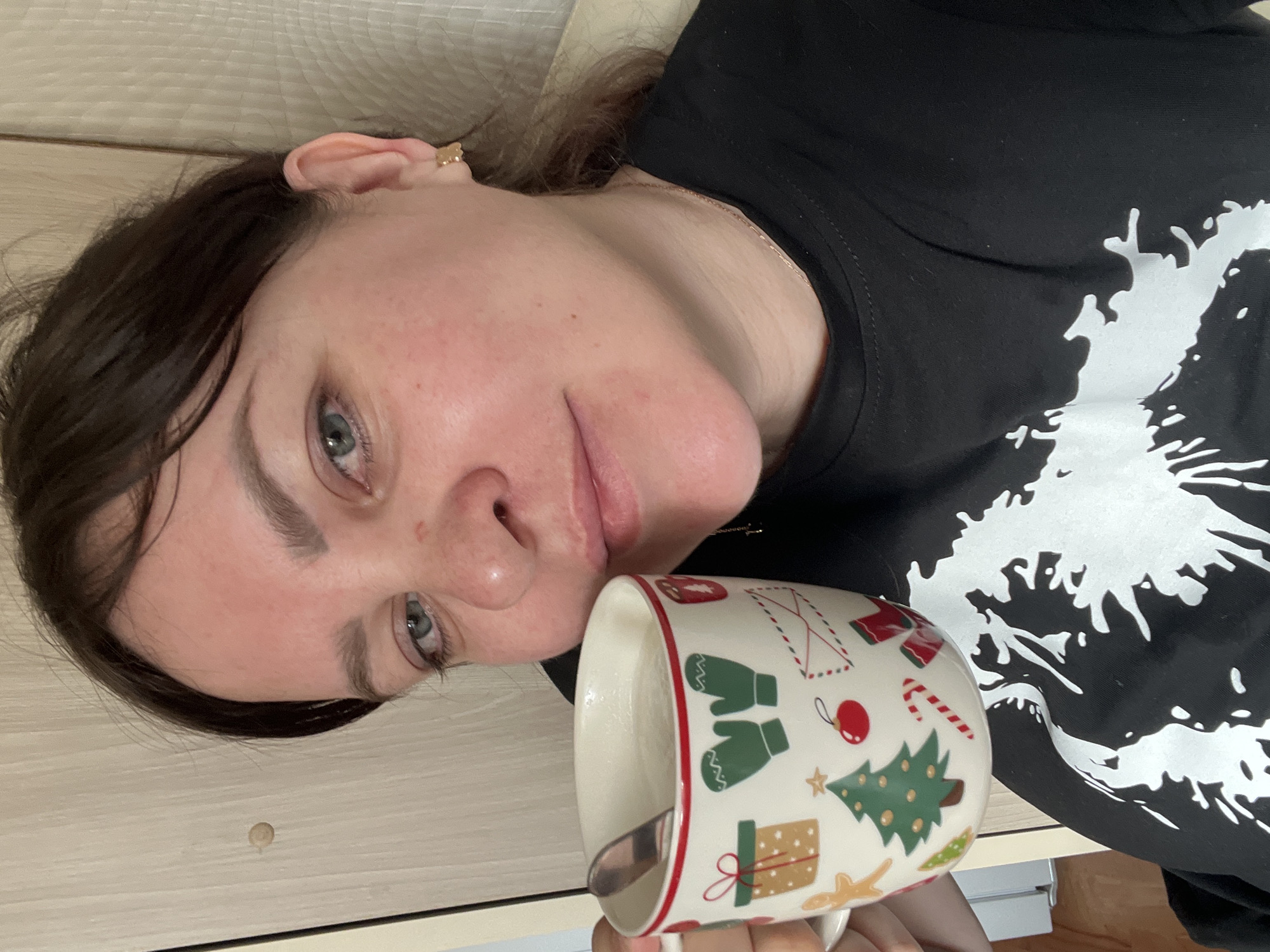 A woman is holding a mug with a spoon in it. The mug is decorated with festive images such as a Christmas tree, a candy cane, and a wrapped gift. The woman is wearing a black shirt with a white print. She has an earring in her left ear. The background appears to be a wall made of light-colored wood panels. The image was most likely taken indoors. There is no indication of the location of Perm, Russia in this image.