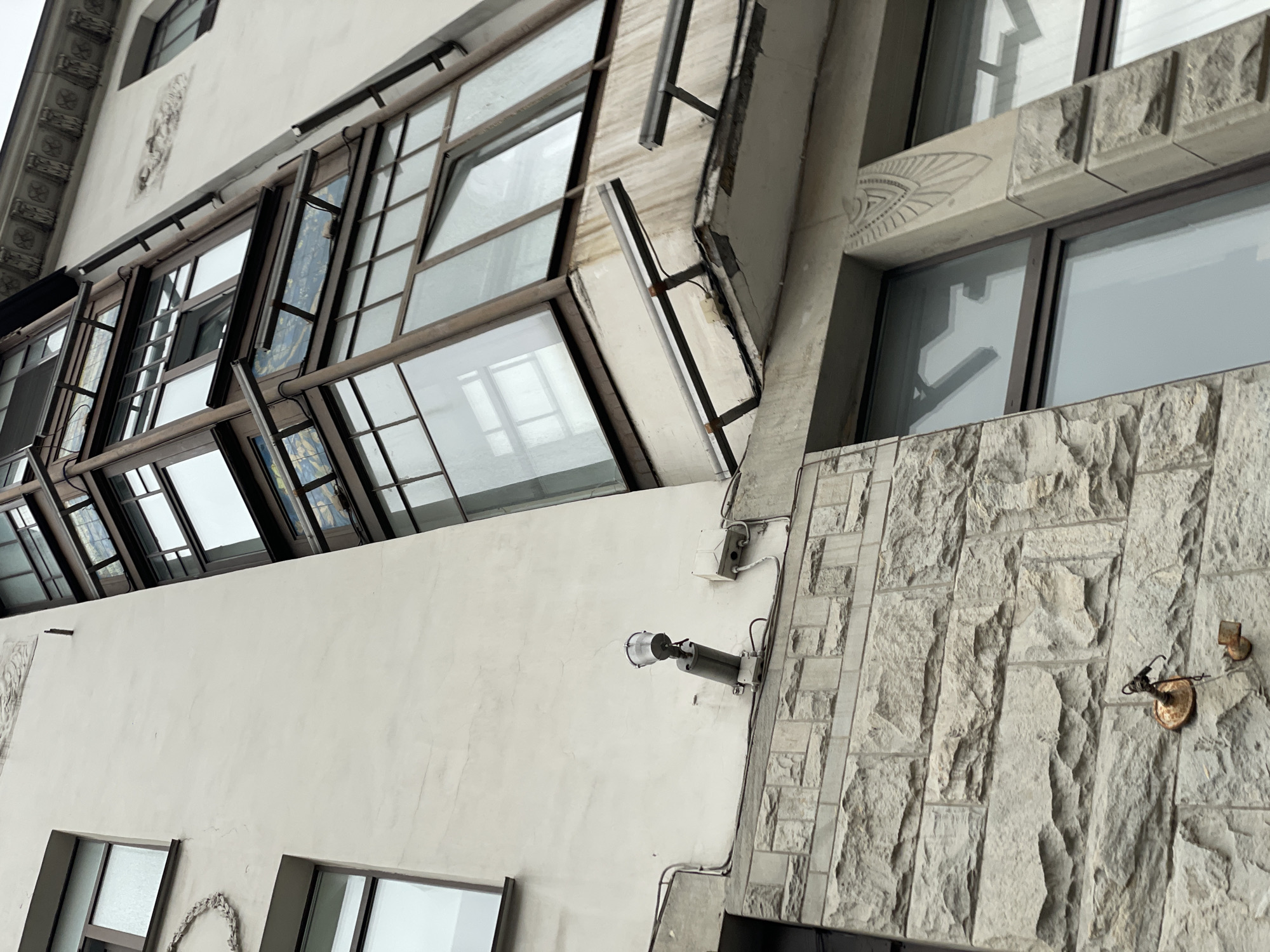 The image depicts a building facade in Saint Petersburg, Russia, exhibiting a mix of architectural styles. The lower portion features a white wall with two visible windows and decorative elements. A surveillance camera is mounted on the wall near the intersection of the white facade and a textured, light-grey stone-clad section. The stone section also features an antiquated metal fixture. Above, a series of windows are arranged in a grid-like pattern with some open at an angle. The building's style appears to be a fusion of classical and modern elements.