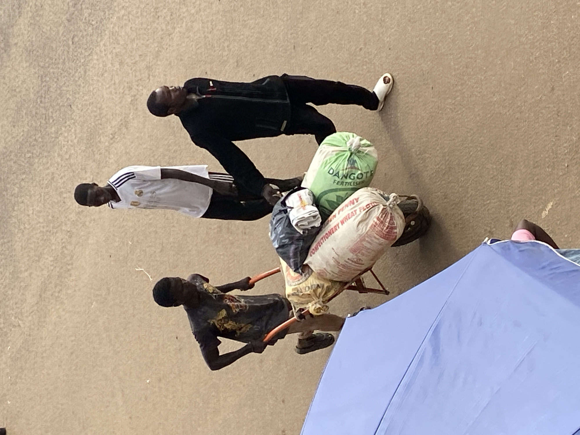 The image displays an outdoor scene in Bukuru, Nigeria, from an elevated perspective. The ground is a light brown, textured surface. In the lower left foreground, a section of a blue umbrella with a white edge is visible. Three dark-skinned males are present.
On the left, one man, wearing a dark short-sleeved t-shirt with a yellow design, dark shorts, and dark shoes, is actively pushing an orange-framed wheelbarrow. The wheelbarrow is loaded with multiple large bags. Visible amongst the load are an off-white bag with red lettering, partially identifiable as "CONFECTIONERY WHEAT FLOUR" and "PENNY," and a green bag with white text indicating "DANGOTE FERTILIZER." Additional items, including a dark bundle and lighter fabrics, are stacked on top of the bags.
Walking alongside the right side of the loaded wheelbarrow is a second man, dressed in a black long-sleeved zipped jacket, dark trousers, and white slip-on sandals. His left hand is positioned near the bags on the wheelbarrow.
Further in the background, slightly to the left of the second man, a third man stands. He is wearing a white short-sleeved t-shirt with black stripes on the shoulders and dark trousers. All three individuals have short dark hair.