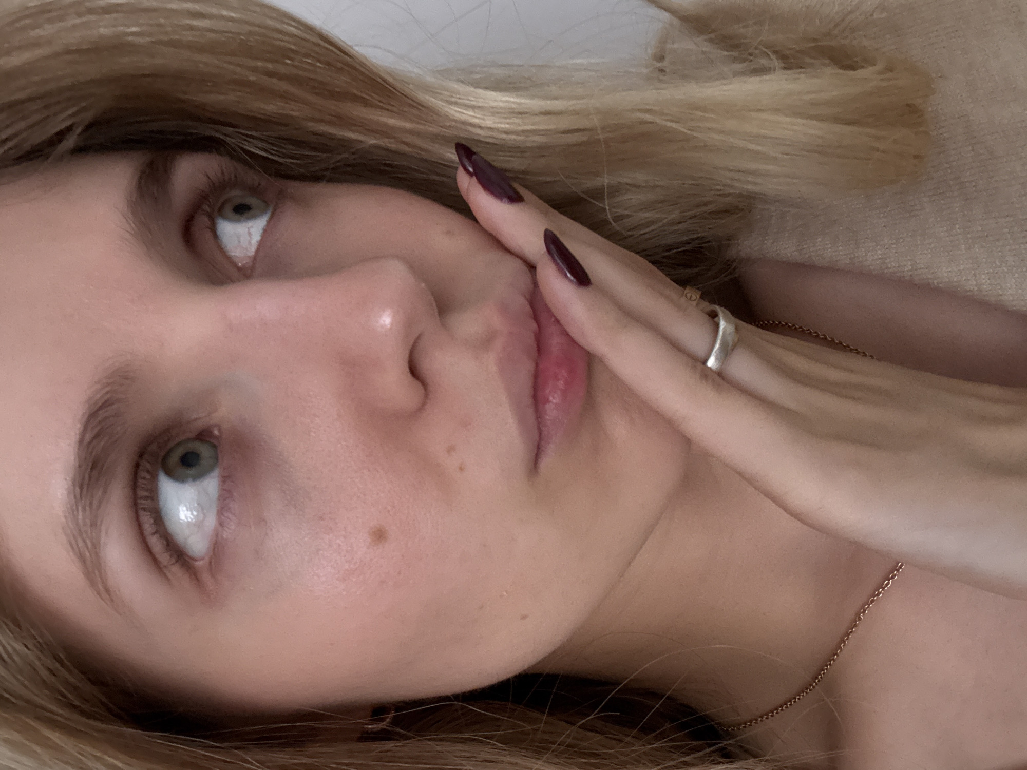 A close-up, tilted view of a fair-skinned person with blonde hair. The person's right hand, adorned with long, dark-colored nails and a silver ring, is touching their face near their mouth. They are lying down, and a gold chain is visible around their neck. The person has light green eyes that are looking upwards. No specific location in Mexico City is discernible from the image.