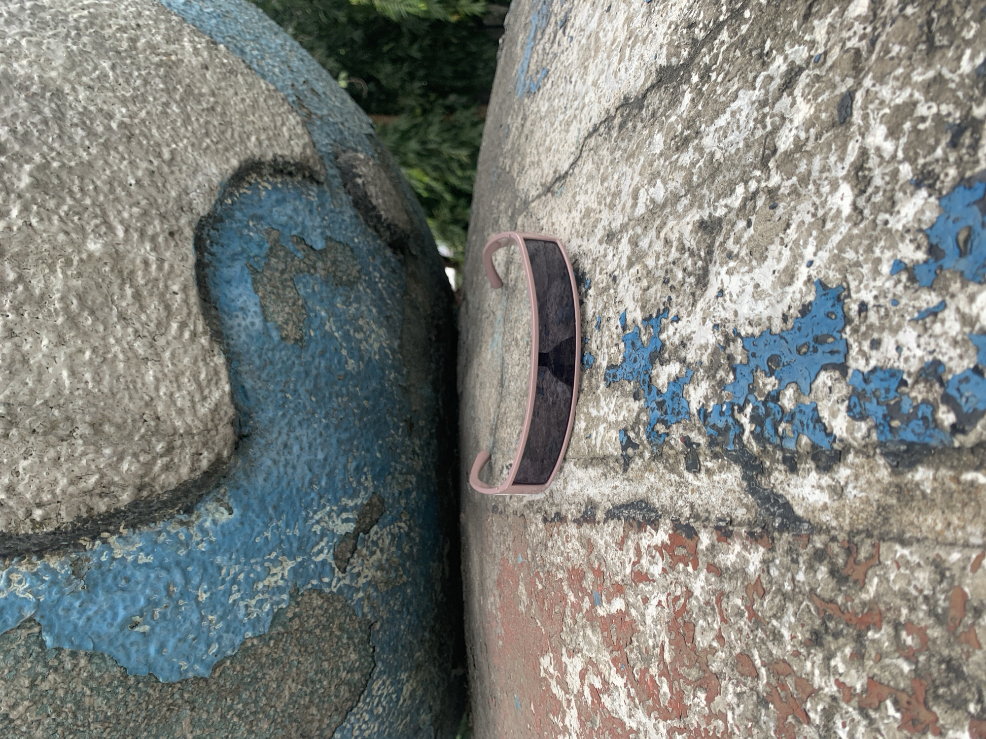 A pair of pink-rimmed sunglasses with dark lenses lies on a weathered, textured concrete surface. The concrete is partially covered with peeling paint in patches of red and blue. In the background, a large, curved, weathered surface with patches of blue paint is visible, casting a shadow over the sunglasses. Green foliage is visible in the upper right background. The scene appears to be an outdoor setting, possibly an urban environment in Iganmu, Nigeria, given the context of the location. There are no people or actions depicted in the image.