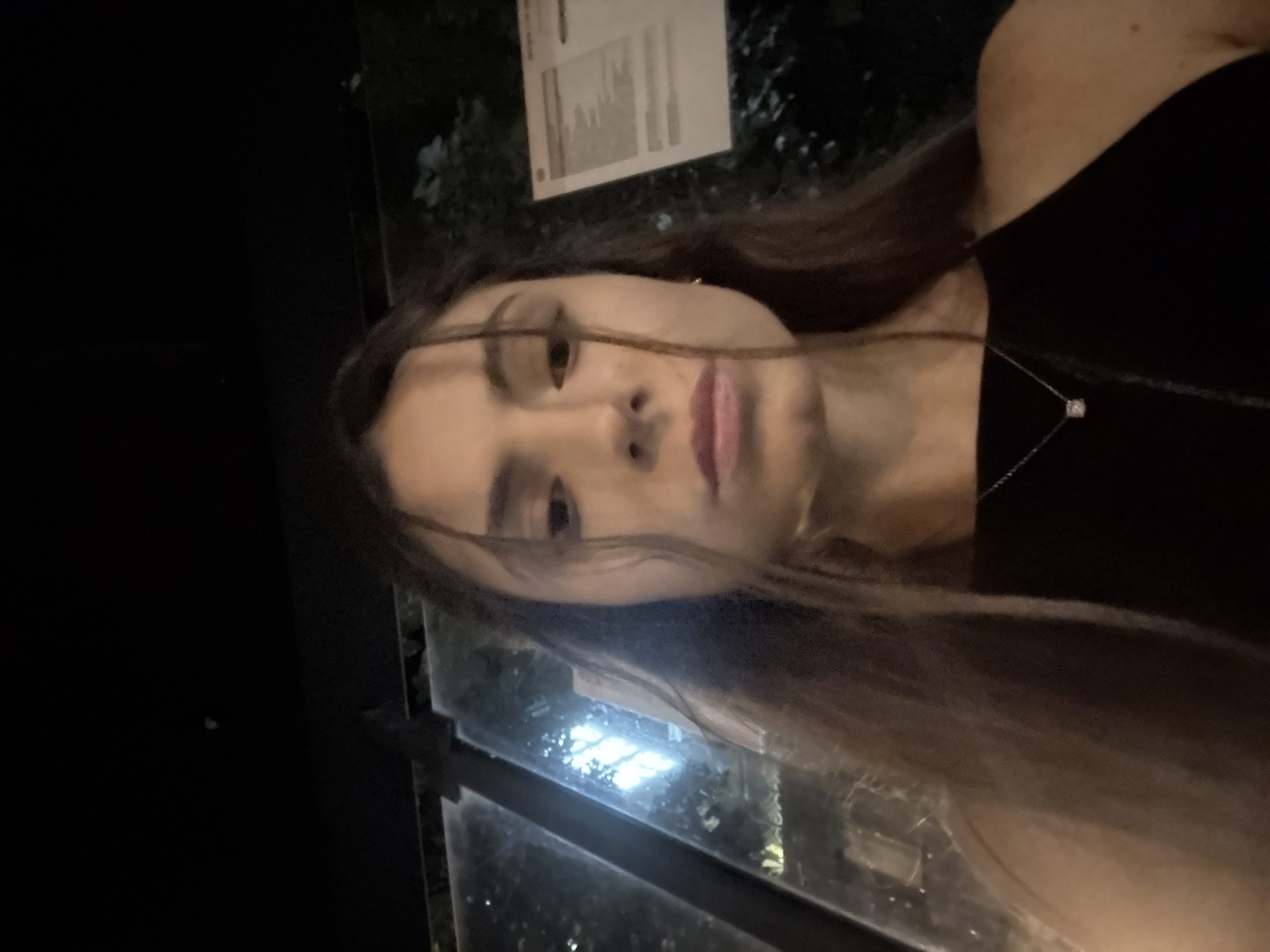 A young woman with long brown hair and dark eyes is looking directly at the camera. She is wearing a black sleeveless top and a delicate silver necklace with a square pendant. She appears to be standing outdoors at night, possibly at a bus stop. The background shows a glass enclosure with reflections of lights, and some foliage can be seen behind the glass. A white sign with text is also visible on the right side of the frame. The scene is dimly lit, suggesting it is nighttime. The location is the city of Varna, Bulgaria.