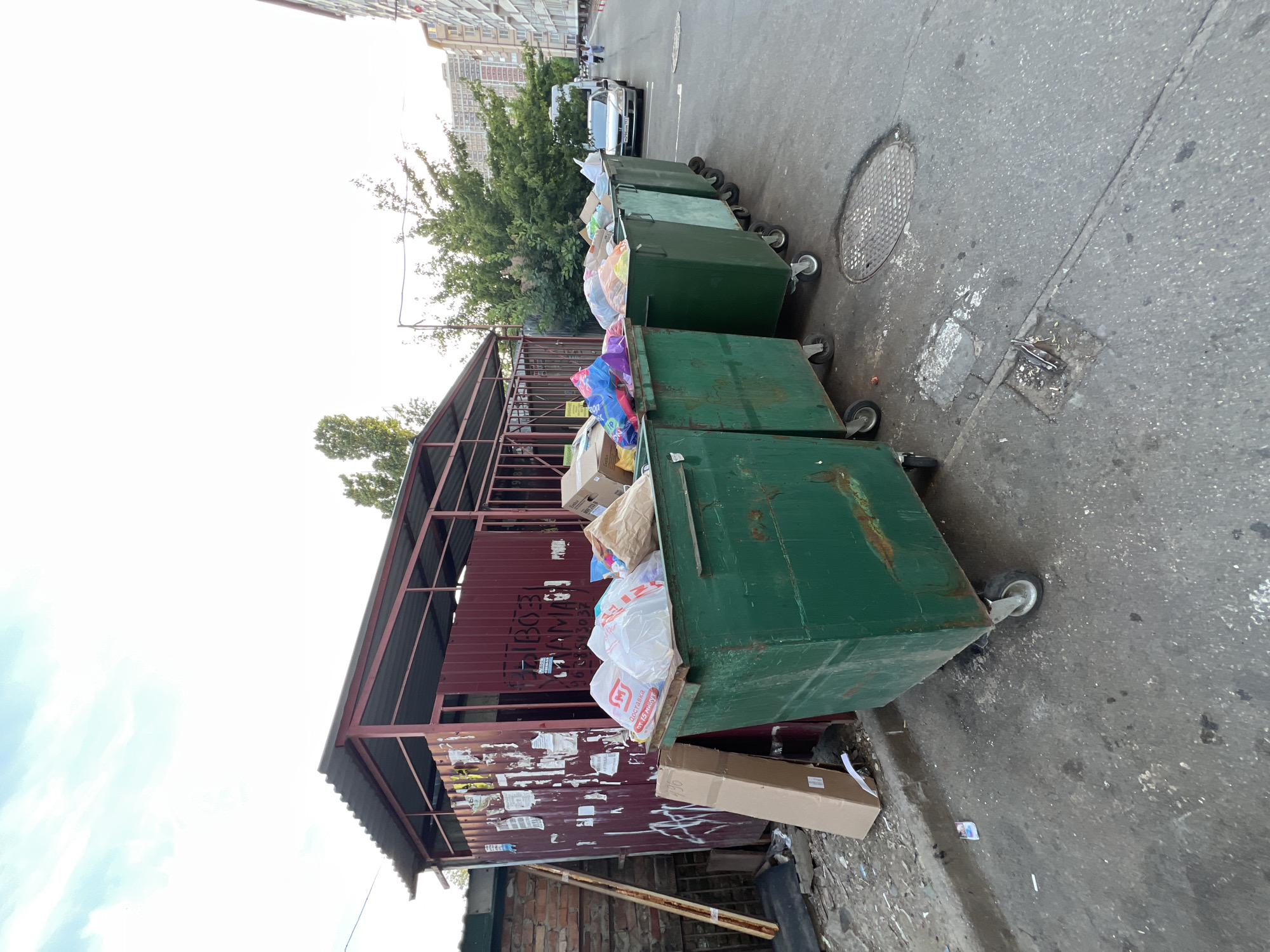 A row of overflowing green dumpsters stands on an asphalt surface in front of a weathered, red metal shelter. The shelter has a corrugated metal roof and its walls are adorned with various peeling posters and graffiti. Cardboard boxes and plastic bags are scattered around the dumpsters and the area. In the background, trees and multi-story residential buildings are visible, suggesting an urban environment. A silver sedan is parked on the right side of the frame, and two people are walking in the distance. The sky is overcast with hints of blue. The scene appears to be a service area for waste disposal in a residential district.