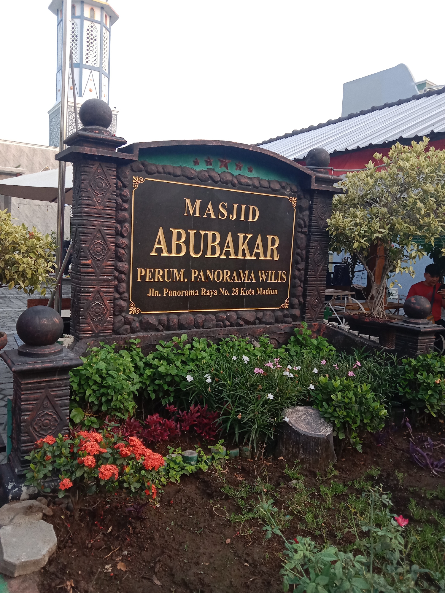 This is a medium-shot, eye-level, outdoor photograph of the entrance sign to Masjid Abubakar, located in Perum. Panorama Wilis, Jl. Panorama Raya No. 28, Kota Madiun, Indonesia. The sign itself is large and black with gold lettering that reads "MASJID ABUBAKAR" on the top two lines, and below that "PERUM. PANORAMA WILIS" and "Jln. Panorama Raya No. 28 Kota Madiun." The sign is framed by a dark, ornate pillar structure with decorative carvings and a green, rocky border.
In front of the sign, a garden bed is filled with lush greenery, including clusters of bright orange ixora flowers and smaller pink and white flowers. A tree stump is visible to the right of the sign, surrounded by more plants. Behind the garden and the sign, a minaret of a mosque can be seen to the left, featuring a white and blue geometric design. To the right of the sign, part of the mosque's roof structure is visible, as is a tree with light green leaves. A person in a red shirt is partially visible in the background on the far right, looking at a phone. The ground is composed of dark soil and some paving stones. The overall lighting suggests it is either early morning or late afternoon.