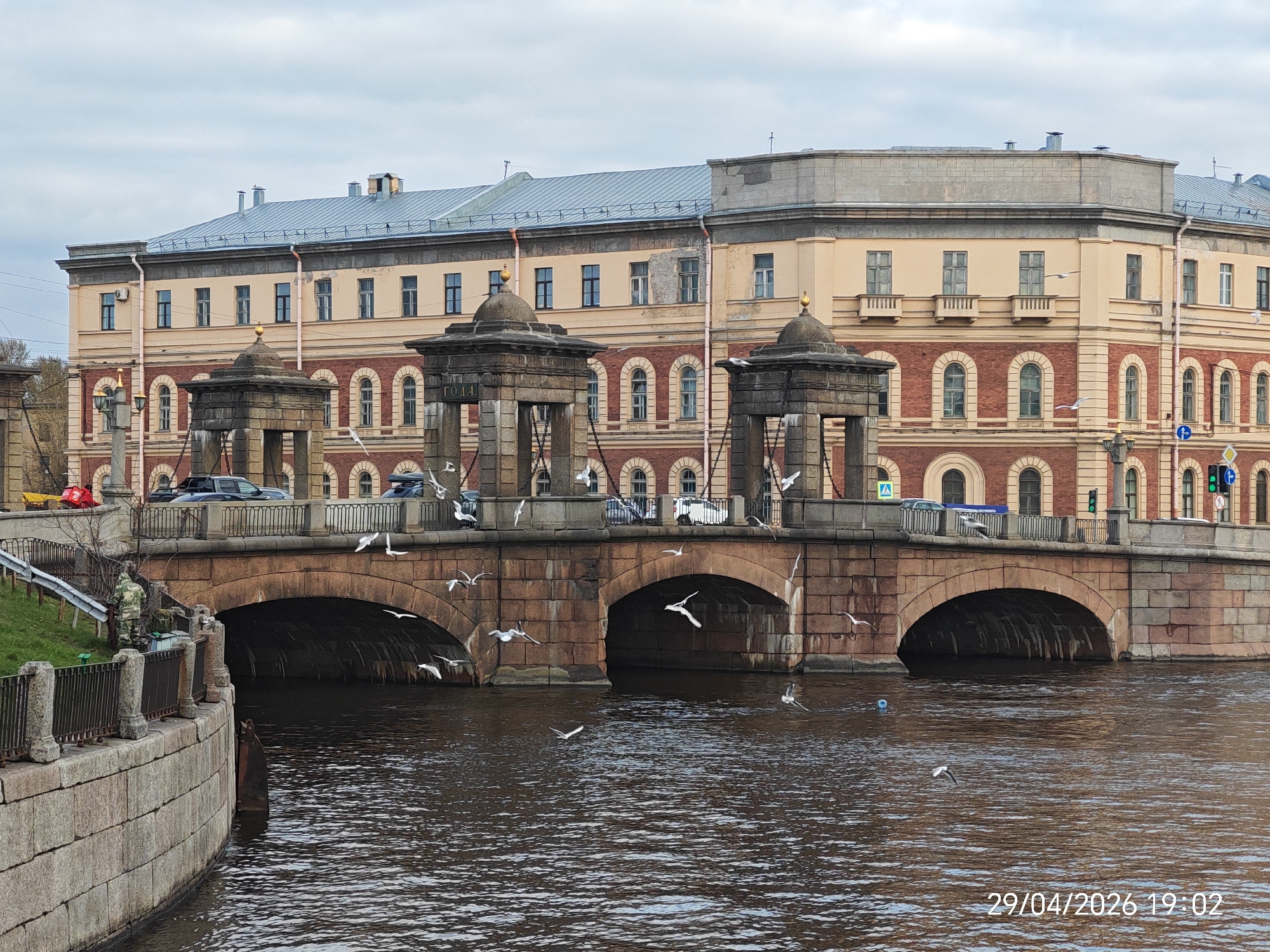 An outdoor photograph captures a stone bridge spanning a dark, rippling canal or river under an overcast sky. In the background, a large, multi-story building features a light yellow upper facade, a red brick lower section with arched windows, and a light blue metallic roof.
The bridge, constructed from large, brown and reddish-brown stone blocks, has at least three arches visible over the water. On the bridge deck, traffic appears to be moving, with several cars visible, including a dark SUV/truck. Four ornate stone pavilions, topped with domes (two with golden finials), are positioned along the bridge, adding a distinctive architectural element.
Numerous white seagulls are seen flying low over the dark water, some with wings outstretched, others closer to the surface, suggesting activity like foraging or communal flight. On the left bank of the canal, a person dressed in camouflage stands near the water's edge beside a metal fence, appearing to observe or attend to something.
The overall atmosphere is calm and mundane, characteristic of a cloudy day. A timestamp in the bottom right corner of the original image reads "29/04/2026 19:02," indicating the early evening. Visible text "ГОДА." (GODA.) can be seen on one of the bridge's stone structures, hinting at a Russian-speaking location. Other details include traffic lights showing a green signal on the right, and various railings along the bridge and canal bank.