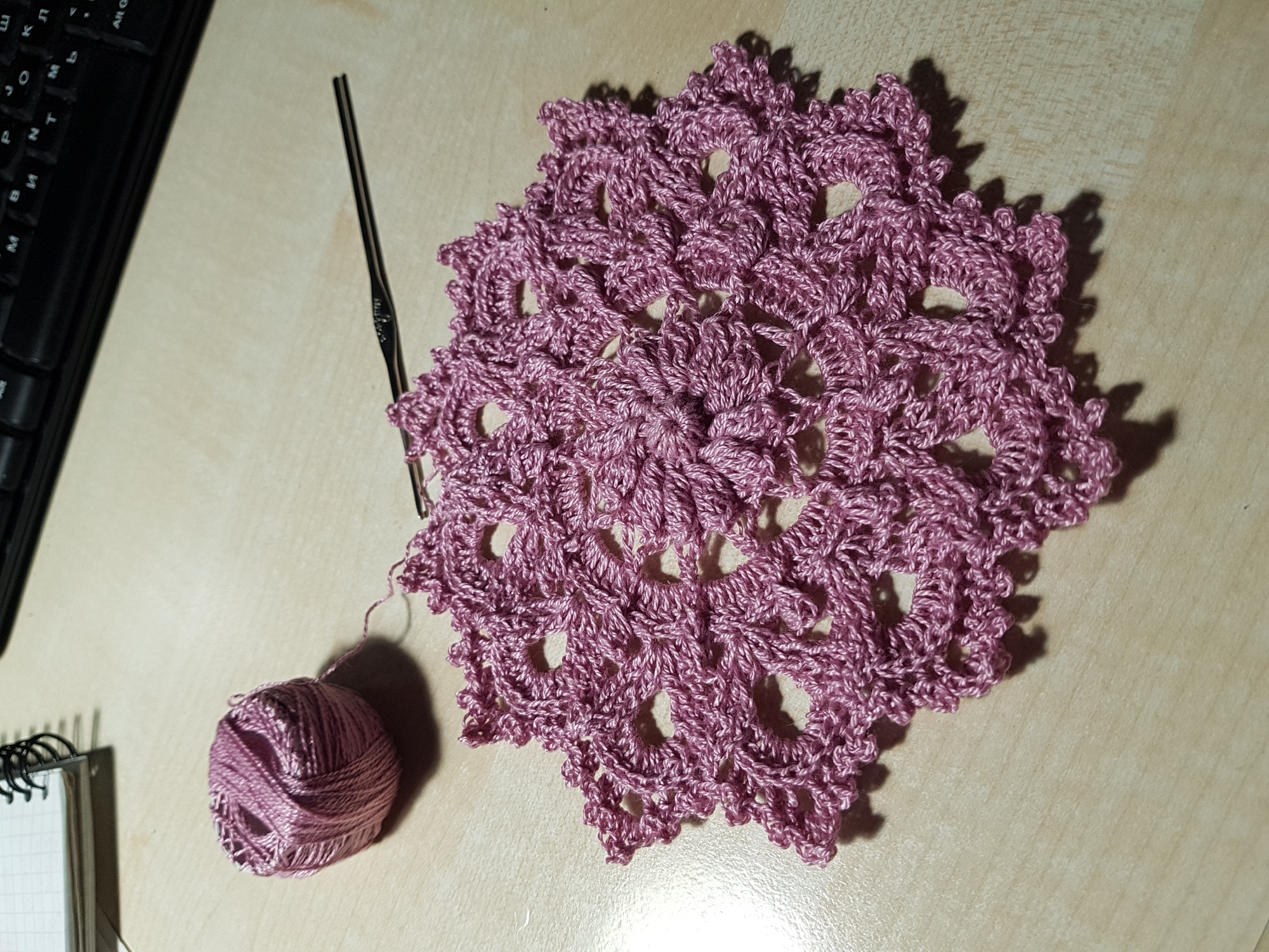 The image depicts a crocheted doily on a light-colored surface. The doily is round with a complex, lace-like pattern and a mauve/pink color. A crochet hook and a ball of matching yarn are positioned near the doily. To the left of the doily is a partially visible computer keyboard and a notebook. The scene appears to be indoors, possibly on a desk or table. There is no indication of Saint Petersburg, Russia.