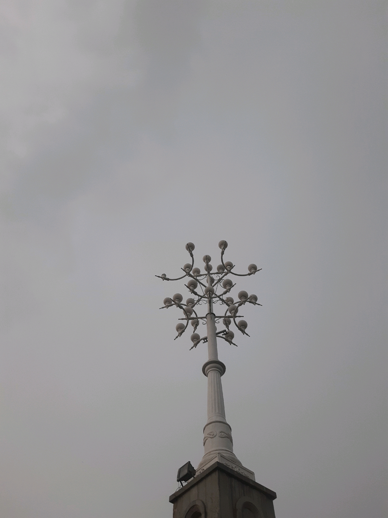 The image captures the upper portion of a decorative lamppost, characterized by its multiple bulb fixtures branching outwards, resembling a stylized tree or candelabra. The lamppost is white and stands tall against a uniformly overcast sky. The base of the lamppost is integrated with a stone structure, possibly part of a building or monument, from which a floodlight is also visible. The scene is set in Tbilisi, Georgia, specifically at the Tbilisi Funicular, located on Mtatsminda Hill. The overall atmosphere is somber due to the cloudy weather.