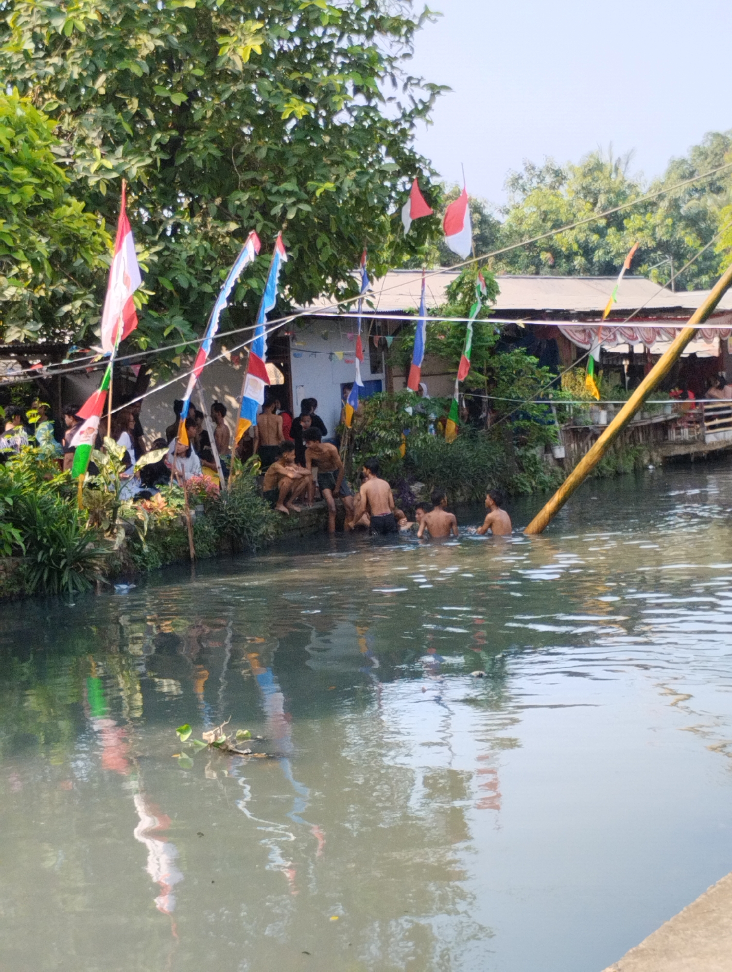 In Bekasi, Indonesia, a group of young men are seen bathing in a murky canal that runs alongside a dense residential area. The canal banks are lined with lush vegetation and cluttered with makeshift structures and some debris, including plastic waste. Colorful flags, including the Indonesian national flag (red and white), are strung across the canal, suggesting a local celebration or event. The water's surface reflects the surrounding trees and buildings, indicating overcast or subdued lighting conditions. Some individuals are fully submerged, while others are partially in the water, with their torsos visible. The scene depicts a common sight in some urban areas of Indonesia where natural waterways are utilized for recreation despite the water quality.