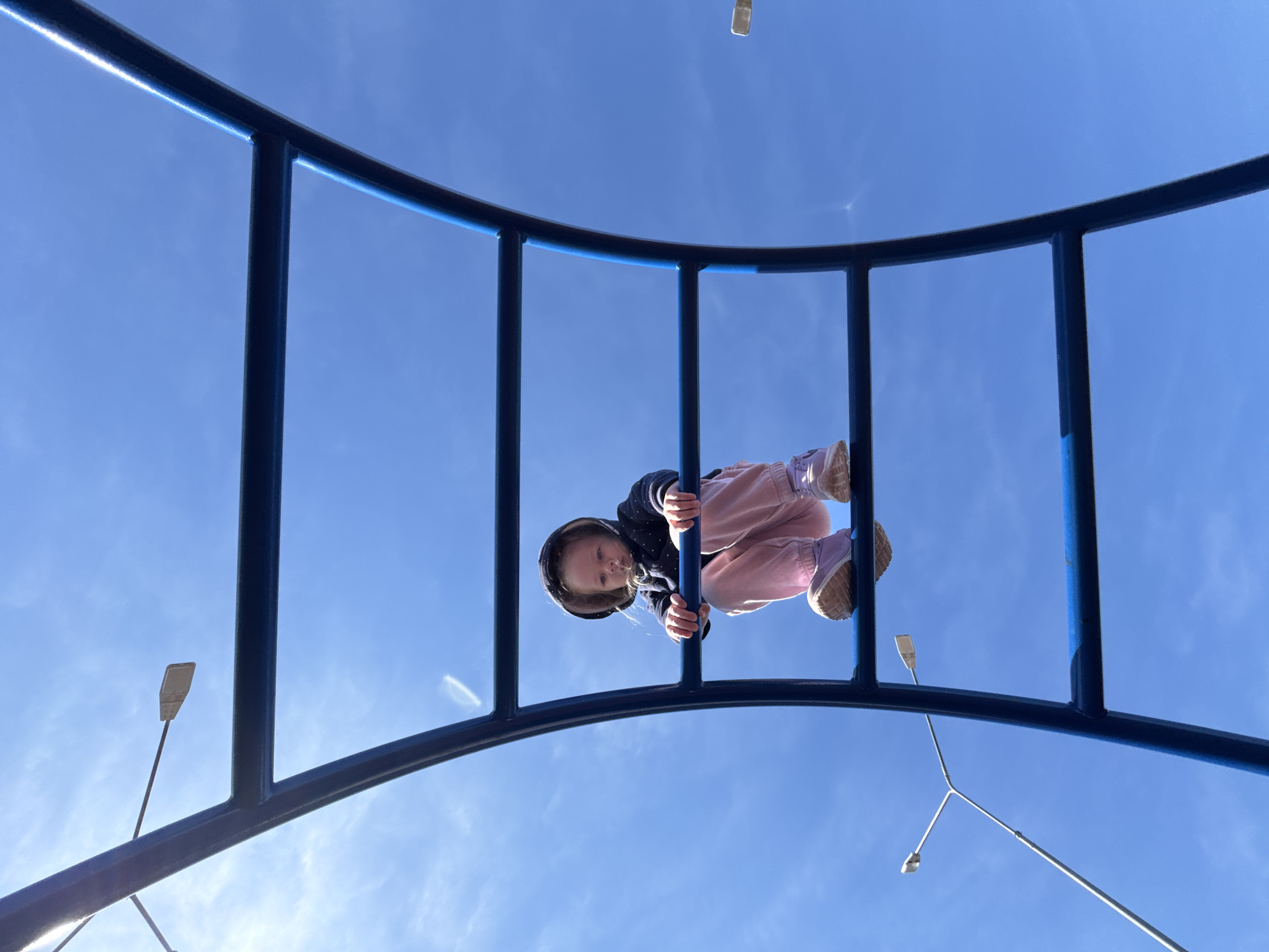 A child is hanging upside down from a blue metal jungle gym. The child is wearing a dark jacket with a hood, pink pants, and light-colored shoes. The jungle gym is set against a clear, blue sky. In the background, street lights are visible. The location is Горный, Russia.