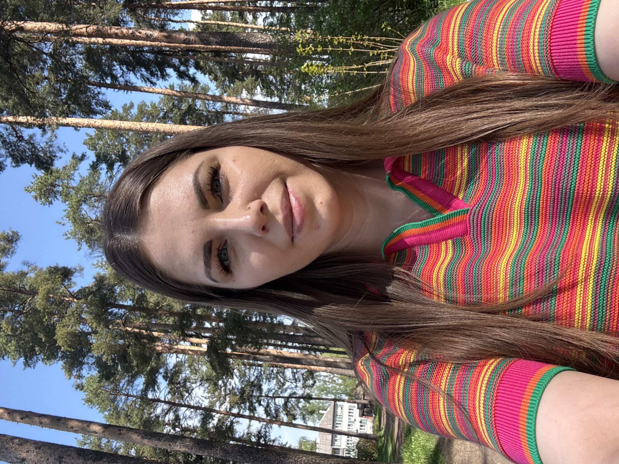 The image shows a woman with long brown hair wearing a colorful striped top, likely a sweater or blouse. The setting appears to be outdoors in a wooded area, with tall pine trees visible in the background against a blue sky. In the distant background, a building is observed. The photo appears to be taken in Razdolye, Russia.