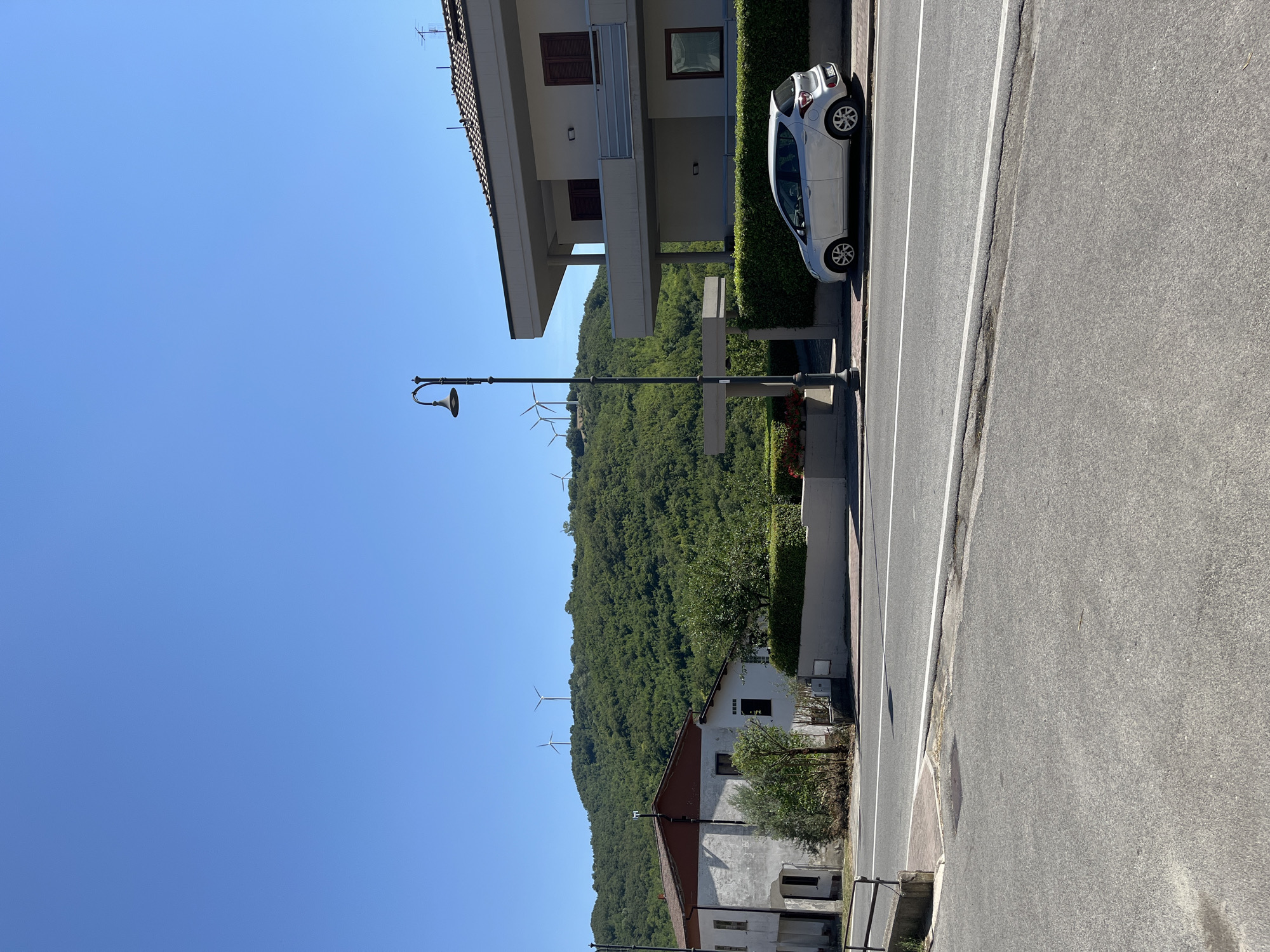 The image depicts a street scene in Foiano di Val Fortore, Italy, under a clear blue sky. On the left, an aged, white building with a reddish-brown roof is visible. Adjacent to it, a modern, multi-story building with a light-colored facade and dark-shuttered windows stands on the right. A silver car is parked on the sidewalk in front of the modern building. A street lamp with an ornate design is positioned between the two buildings, near the sidewalk. The background features a lush, green, tree-covered hillside with several wind turbines dotting the summit. The foreground shows a paved road with white lane markings.