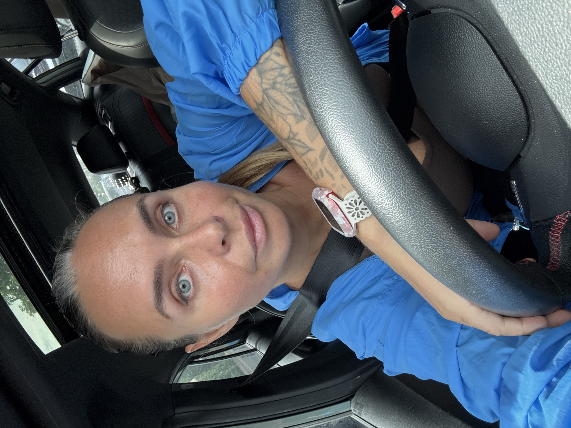 A woman with blue eyes and blonde hair, wearing a blue jacket and a smartwatch, is in the driver's seat of a car. She is holding the steering wheel with her left hand, which has a tattoo on her arm. The car has a black interior with a sunroof. The scene is likely taking place during the day, as there is natural light coming from the sunroof. The location is not identifiable as Bangkok, Thailand, or any specific city.