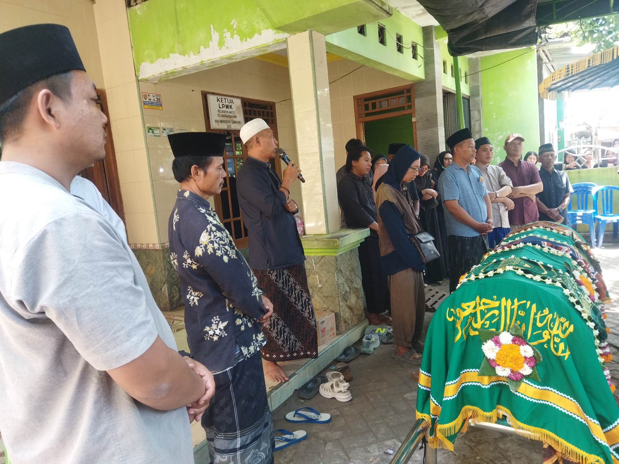 In a courtyard setting likely in Semarang, Indonesia, a group of people, predominantly men wearing traditional Islamic attire including peci caps and sarongs, are gathered around a coffin covered with a green cloth adorned with yellow trim and Islamic calligraphy. A man in the foreground is seen from the back, wearing a light gray shirt and a black peci. To his right, another man in a dark patterned shirt and sarong stands with his hands clasped. A man in the middle is addressing the group while holding a microphone. To the right of the coffin, several women are partially visible, dressed in hijabs. The environment features a building with light-colored walls and green painted window frames, indicating a residential or community building, possibly a mosque or a local community center. Several pairs of footwear are scattered on the ground near the entrance of the building. The lighting suggests it is daytime with sunlight casting shadows.