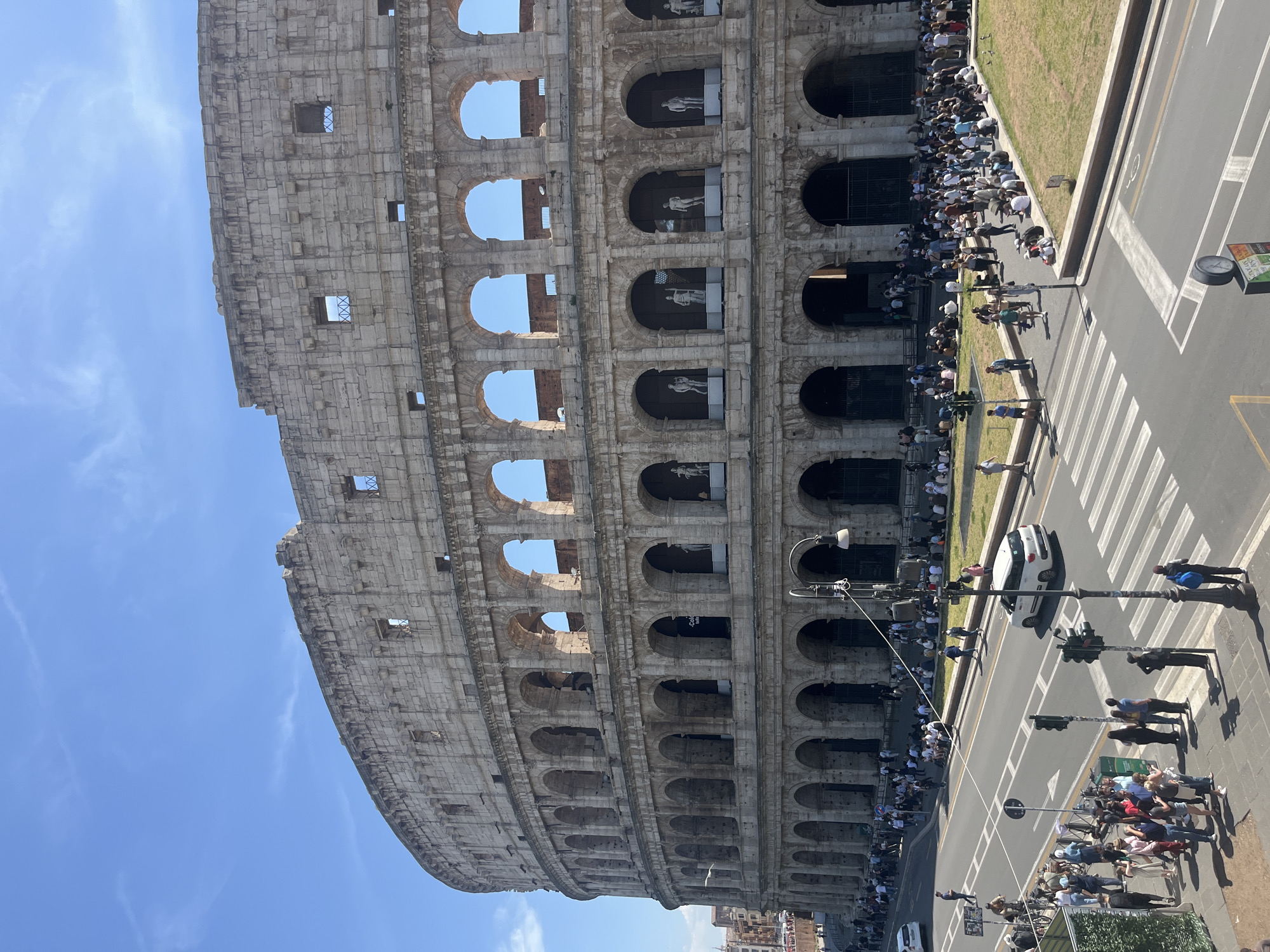 This vibrant daytime image captures the iconic Colosseum in Rome, Italy, from an elevated perspective. The ancient amphitheater dominates the left side and top of the frame, showcasing its impressive multi-tiered stone facade with numerous arched openings. A long line of people, presumably tourists, can be seen queuing along the base of the Colosseum, stretching far into the distance. Several arches on the upper levels appear to house statues or decorative figures, while others frame the clear blue sky. A small, dark sign visible within one of the Colosseum's arches displays the text "Colosseum."
Separating the Colosseum from the foreground is a bustling street with a prominent white-striped pedestrian crosswalk. Several cars, including a white sedan, are present on the road, alongside numerous pedestrians on the sidewalks and waiting near traffic signals. Streetlights and traffic lights are visible fixtures along the road. The foreground also features a strip of green grass and hints of other urban buildings and structures.
The scene unfolds under a bright, clear blue sky with only a few wispy clouds, indicating sunny weather. The overall impression is one of a busy tourist destination, with many people engaged in sightseeing and traversing the area.