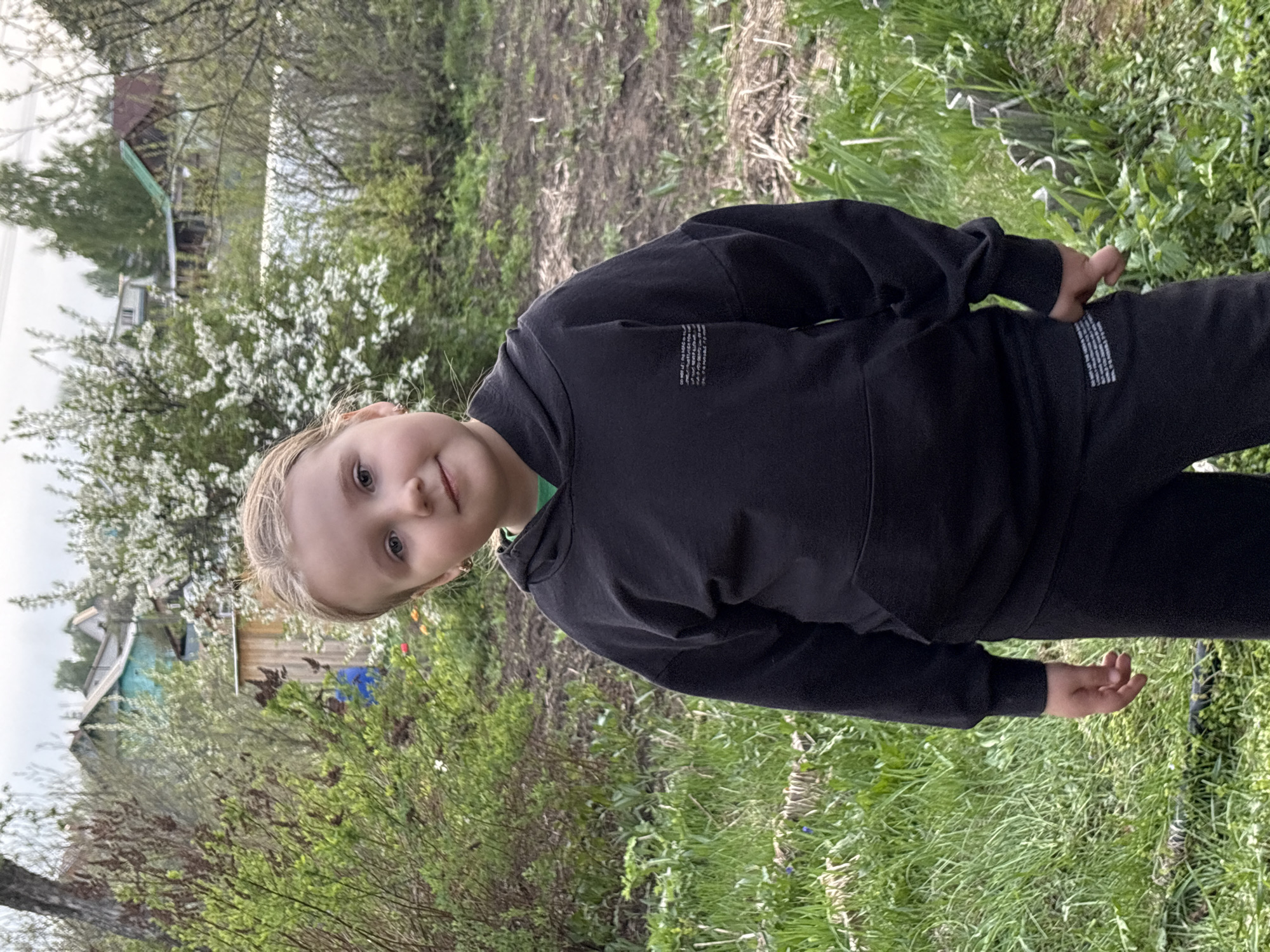 A young, fair-skinned child stands outdoors, presumably in the city of Chaikovsky, Russia. The child, wearing a black hooded sweatshirt and matching pants, is positioned in front of a grassy area interspersed with various plants and foliage. Behind the child, there is a building with a blue roof, a blossoming tree with white flowers, and bare trees in the distance. The ground is covered with grass and patches of exposed dirt.