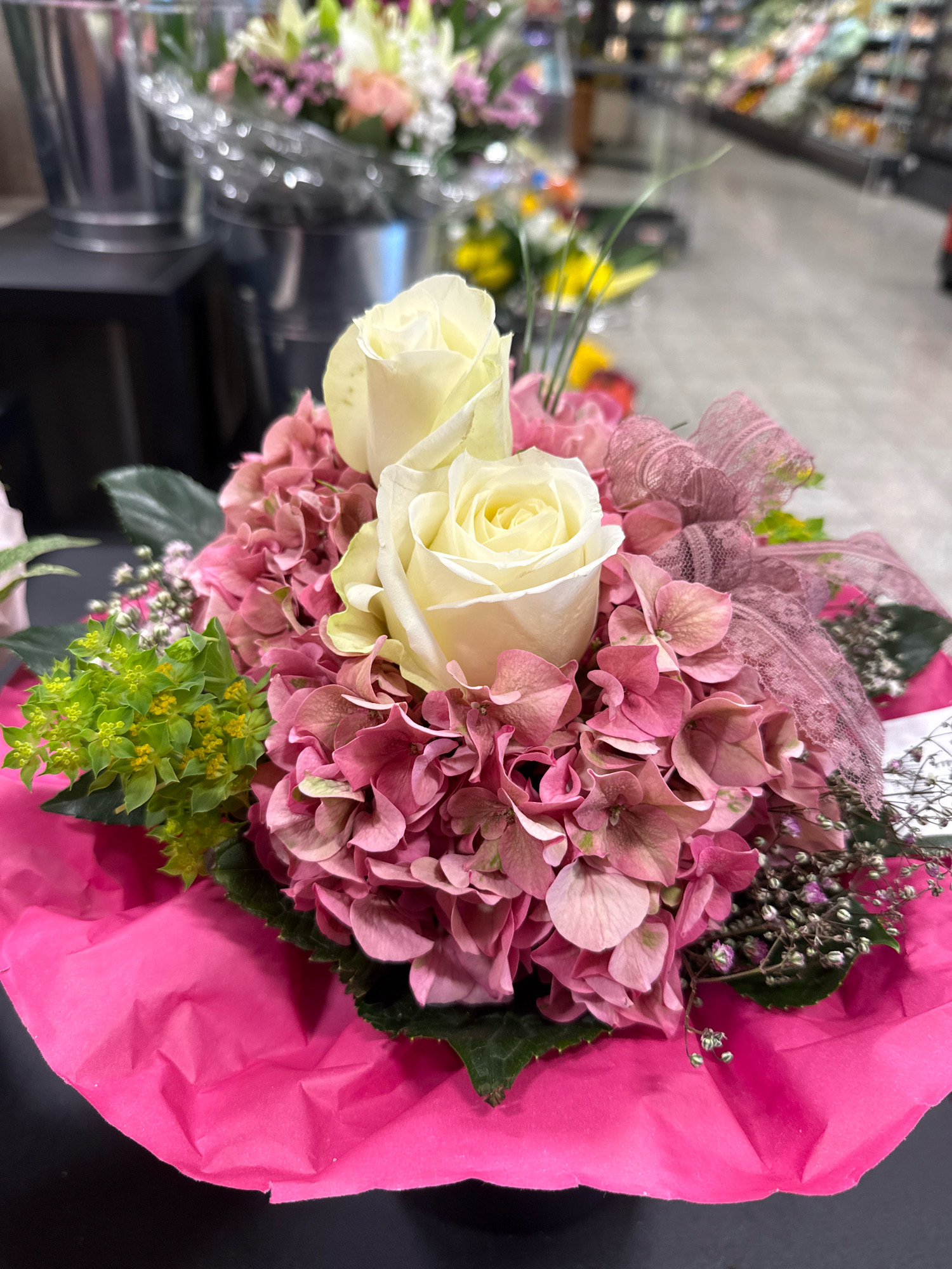 A close-up shot captures a bouquet of flowers, predominantly featuring two cream-colored roses as the focal point. The roses are nestled amongst a bed of pink hydrangeas and accented by small clusters of green foliage with yellow buds and delicate sprigs of tiny white flowers. A sheer pink ribbon with a textured pattern is artfully tied around the bouquet. The arrangement is presented on a surface covered with vibrant pink tissue paper, which drapes over a dark, flat surface, likely a table. The background is blurred, but suggests an indoor environment, possibly a flower shop or market, with shelves of other flowers and metallic buckets visible. This scene is likely located in Bad Lauterberg im Harz, Germany, given the context of the image.