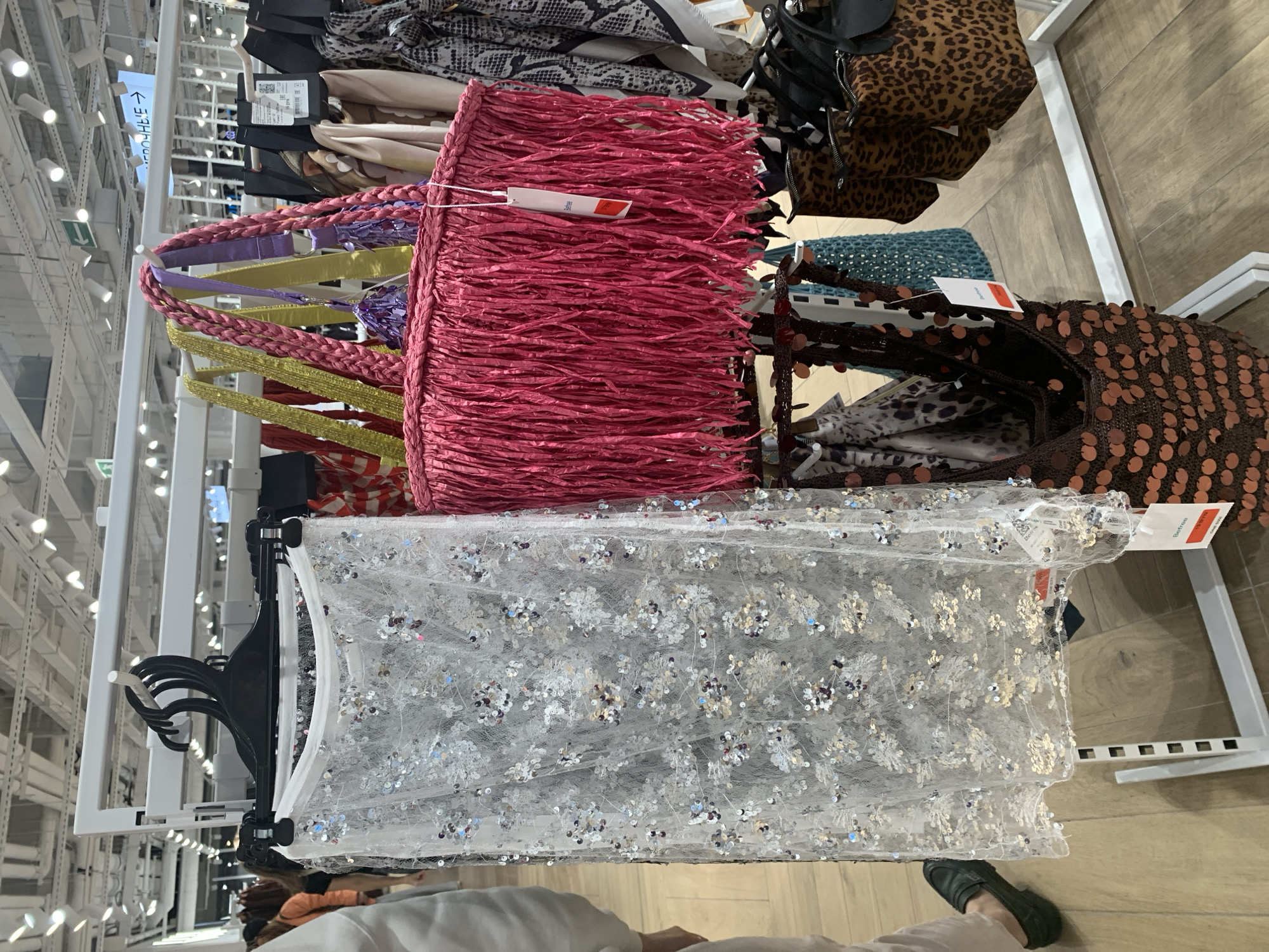 The image shows a retail store display featuring women's fashion accessories. In the foreground, a white, sheer skirt adorned with silver sequins hangs on a white rack. To its right, a large, bright pink woven bag with a fringe detail is displayed. Behind these items, various other bags and scarves with different patterns, including leopard print, are visible. The background reveals a brightly lit store interior with rows of clothing racks and ceiling lights. The floor is made of light-colored wood. In the lower left corner, a person's legs and feet, wearing dark loafers, are visible, indicating the presence of a shopper. The retail environment suggests a commercial setting, likely within a shopping mall or department store. The location is identified as Hrodna, Belarus.