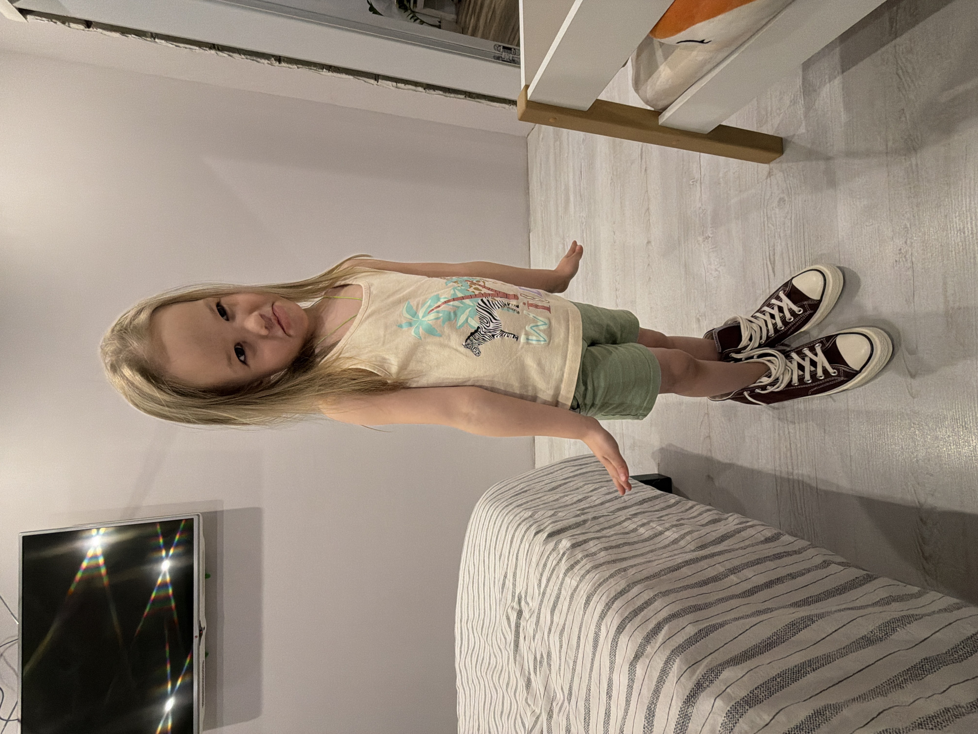A full-body, portrait-oriented photograph depicts a young child, likely a girl, standing indoors in what appears to be a bedroom in Chaikovsky, Russia. The child has long, light blonde hair and is wearing a light cream or yellow tank top with a graphic design featuring a stylized black and white zebra surrounded by green and blue palm trees. She is also dressed in light green shorts and dark brown high-top sneakers with white laces and soles. The child is looking directly at the camera with a slight smile or smirk, her head tilted slightly. Her left hand rests gently on a bed covered with a light-colored, striped duvet.
The room features light-colored wooden or laminate flooring and plain light gray walls. In the bottom left corner, a flat-screen television is mounted on the wall, showing colorful glare patterns on its screen, indicating an indoor light source. In the upper right, part of a white bed frame or bunk bed structure is visible, with a wooden support beam. An orange object, possibly a pillow or stuffed animal, is partially seen behind the white frame. A thin, dark horizontal line runs along the top of the wall, potentially a ceiling trim or structural detail. The image is captured from a slightly low perspective.