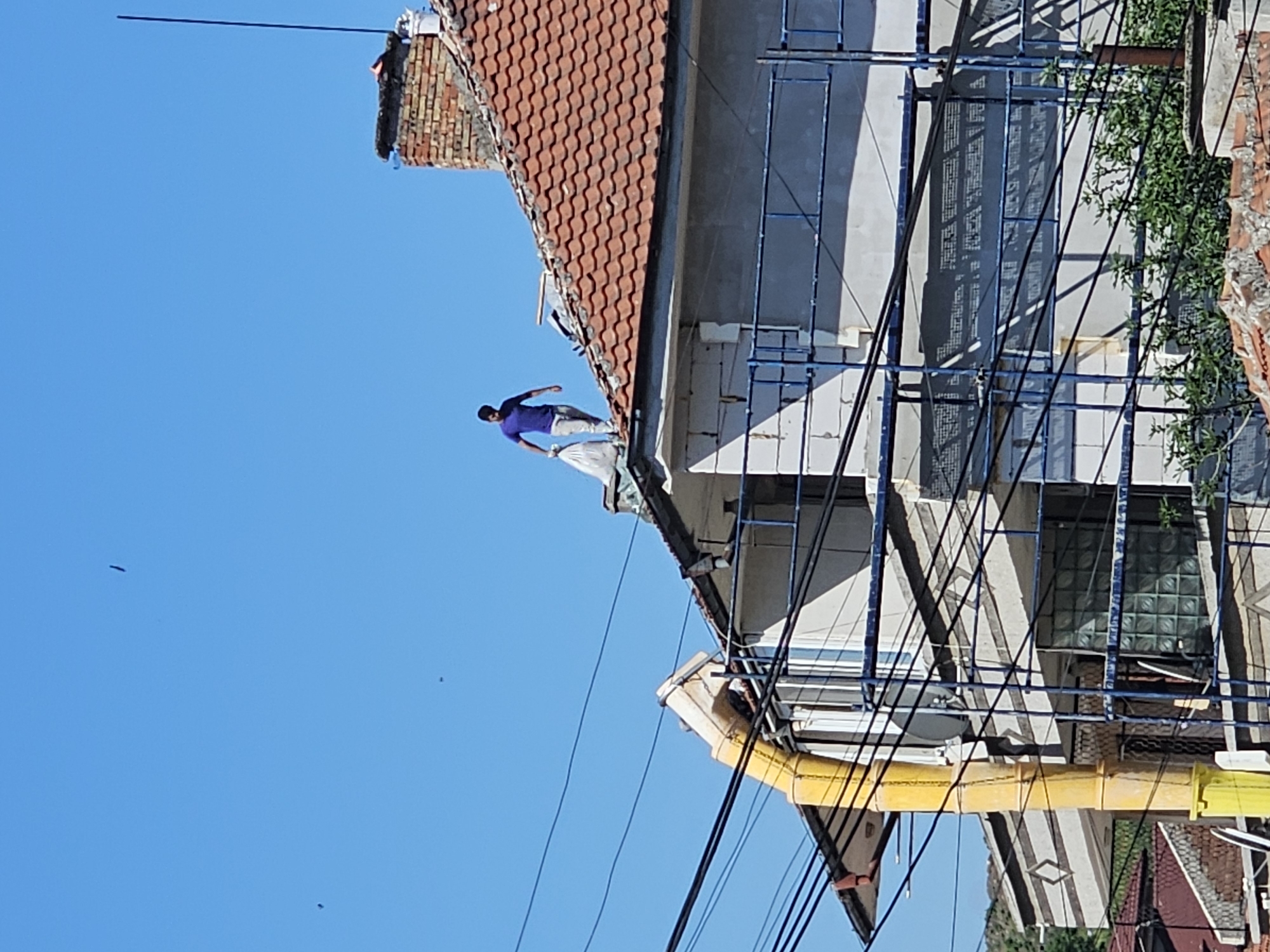 A man in a purple shirt and light-colored pants is walking along the edge of a red-tiled roof of a building in Karnobat, Bulgaria. The building is under construction, with scaffolding visible along the side. Power lines are strung across the lower portion of the frame, and there is a yellow pipe near the building's corner. The sky is clear and blue.