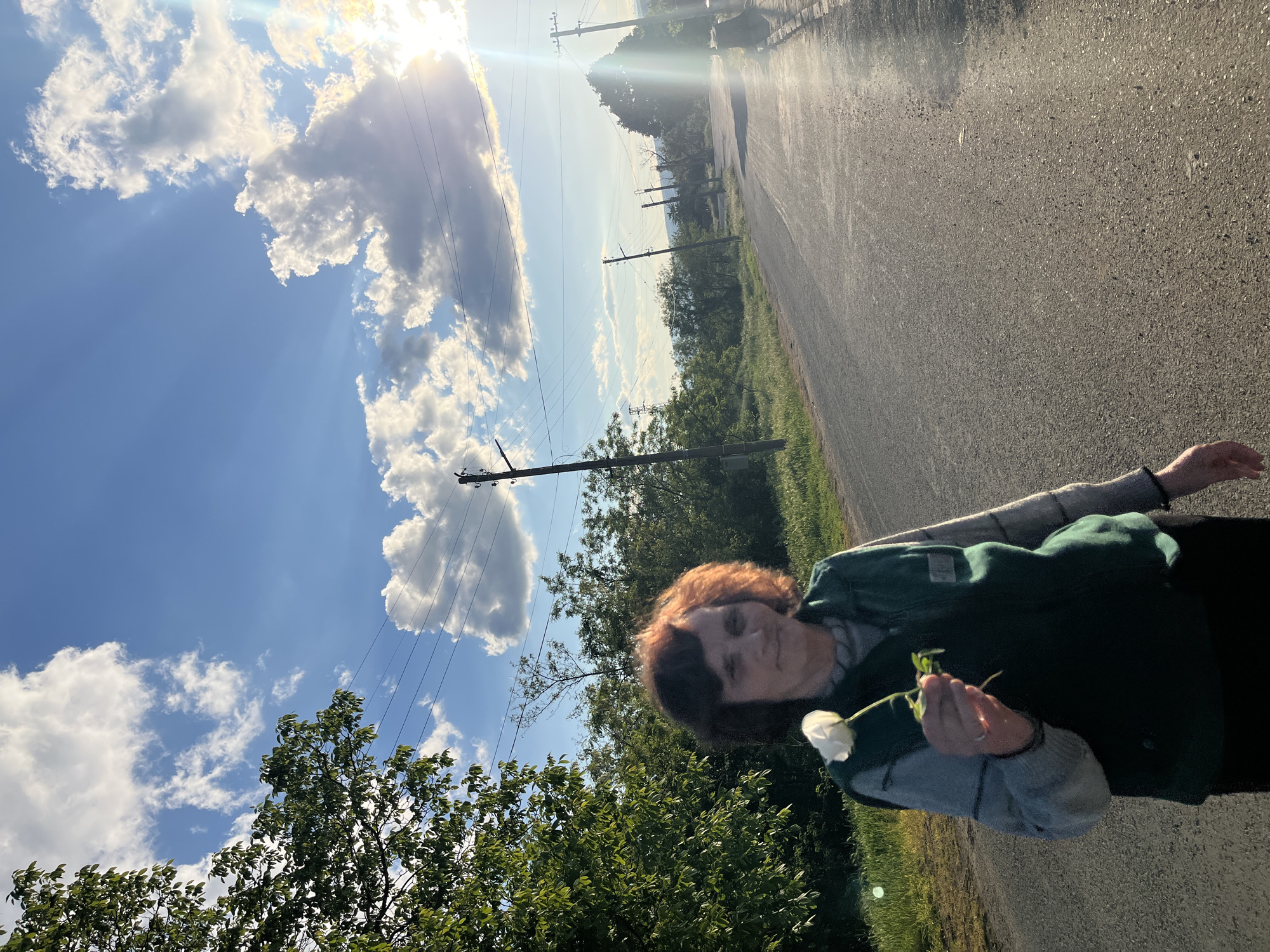 The image depicts a woman holding a white flower, standing by a road on a sunny day. The location is Lozarevo, Bulgaria. The woman has reddish-brown hair and is wearing a dark green jacket over a gray sweater. The sky is blue with fluffy white clouds and the sun is shining brightly, creating lens flare. Power lines are visible in the background. The road is asphalt and there is greenery on either side of it.