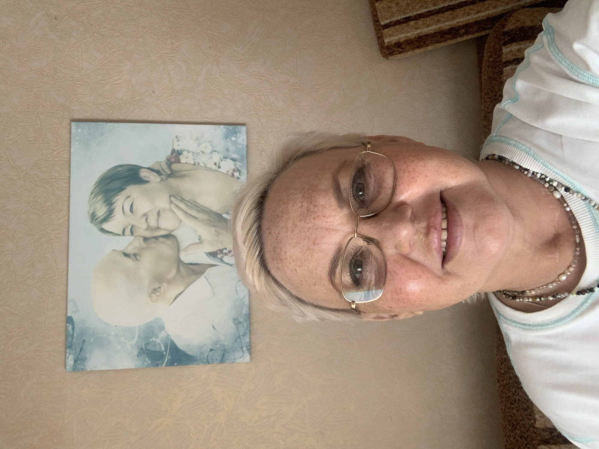 A photograph shows a woman in a white t-shirt and gold-rimmed glasses, smiling at the camera. She is wearing two layered necklaces. To the left of the woman is a framed portrait of a child kissing a bald person. The wall is a beige color. A striped blanket is visible behind the woman's shoulder. The location is not specified.