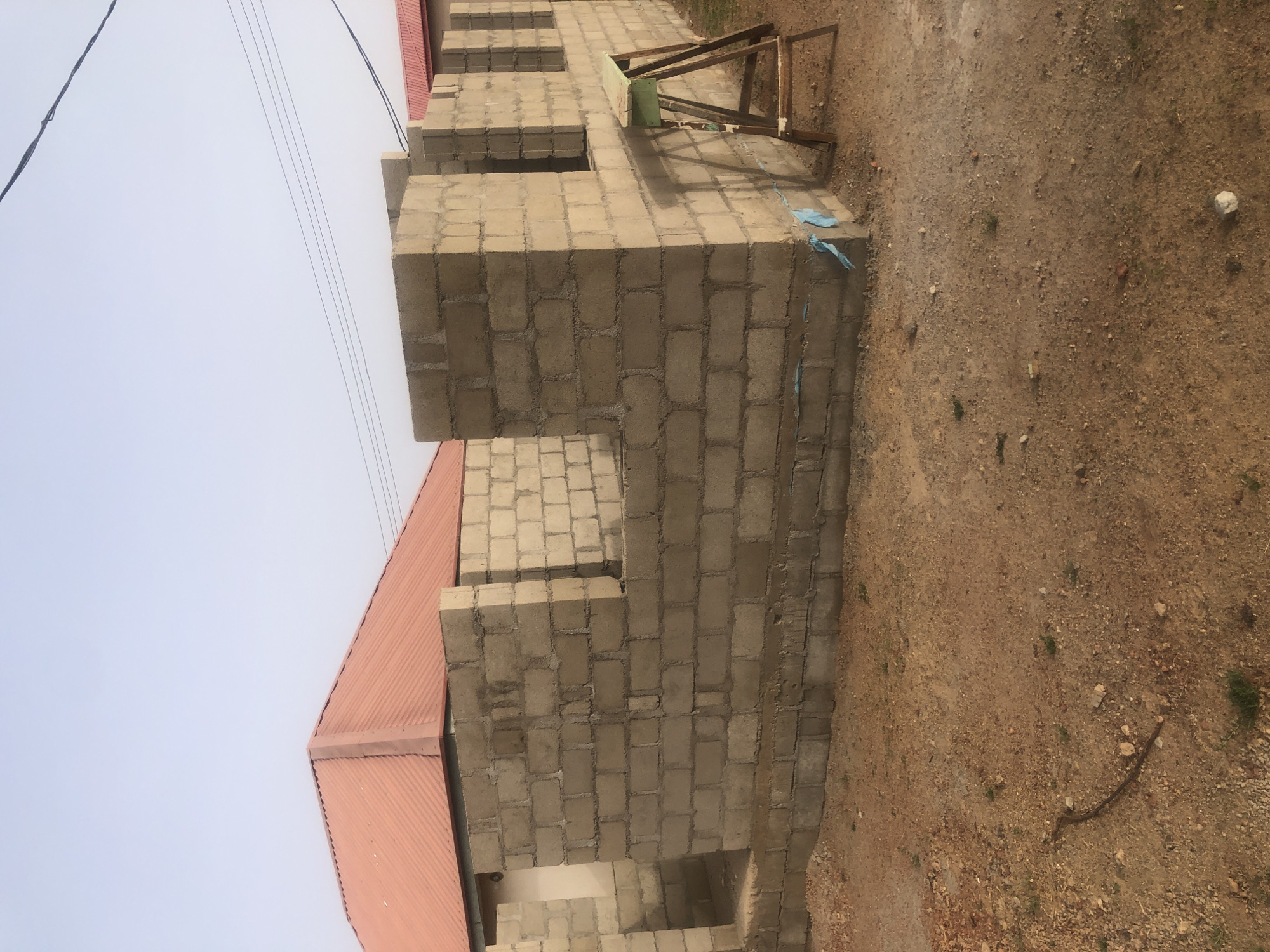 A partially constructed building is visible in a location within Jos, Nigeria. The building is made from gray concrete blocks and a red roof is visible on a part of the building. There is a wooden frame resting on the building. The building is surrounded by dirt and sand. There are electric lines overhead. The sky is a light blue color.