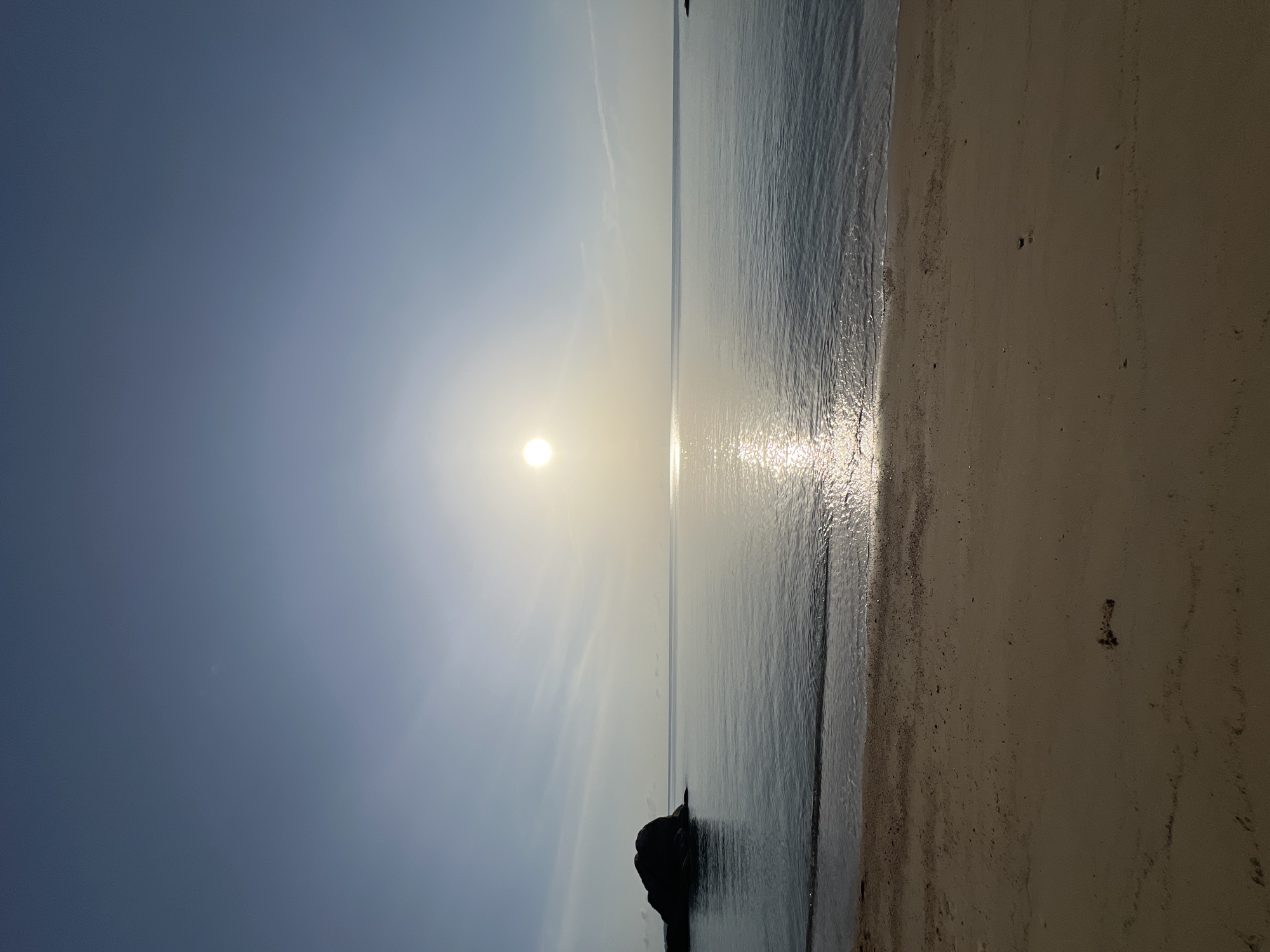 This is a horizontal, full-frame, outdoor, daytime, eye-level shot of a serene beach scene in Ko Samui District, Thailand. The sun is high in the sky, casting a bright glow on the calm ocean water. A small wave is gently lapping at the sandy shore in the foreground. To the left, a large, dark rock formation is partially submerged in the water. The beach sand is a light brown color and appears mostly smooth with some scattered debris. The sky is a clear blue with a few wispy clouds, typical of a sunny day. The overall impression is one of tranquility and natural beauty.