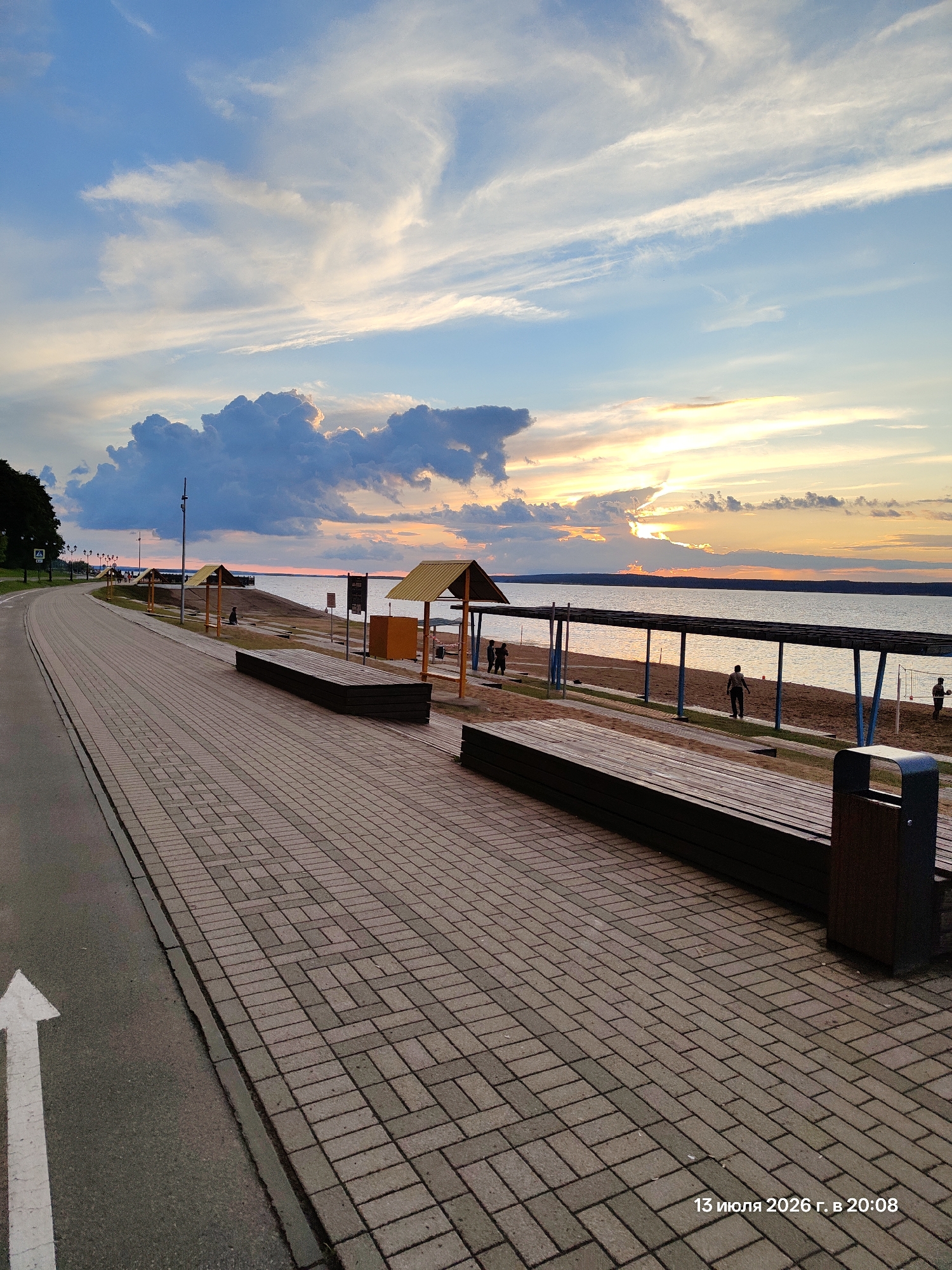 This is a wide-angle, eye-level, outdoor, full shot of a paved promenade alongside a body of water during sunset. The promenade, made of tan and gray interlocking bricks, occupies the foreground and leads to the right. A white arrow is painted on the asphalt road to the left of the promenade. In the mid-ground, there are several wooden benches facing the water. Beyond the benches, there are a few beach umbrellas with yellow roofs and a beach with sand. Some figures are visible on the beach, including what appears to be a volleyball game and individuals walking. The water, a calm expanse of gray-blue, meets the horizon where the sunset is occurring. The sky is filled with a mix of dark blue, gray, and wispy white clouds, with streaks of orange and yellow light from the setting sun peeking through. The overall scene is one of a tranquil evening on a waterfront promenade.