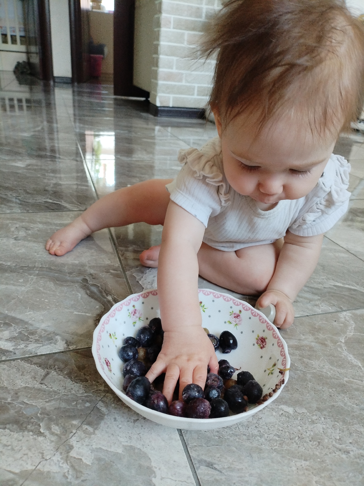 A baby is sitting on a marbled floor, reaching into a bowl of blueberries. The baby is wearing a white shirt with ruffles on the sleeves and is facing down, looking at the blueberries. The bowl is white with a pink floral pattern around the rim. The floor is made of large grey marbled tiles. In the background, there is a doorway and a brick wall. This scene is indoors, likely in a home located in Vinogradny, Russia.