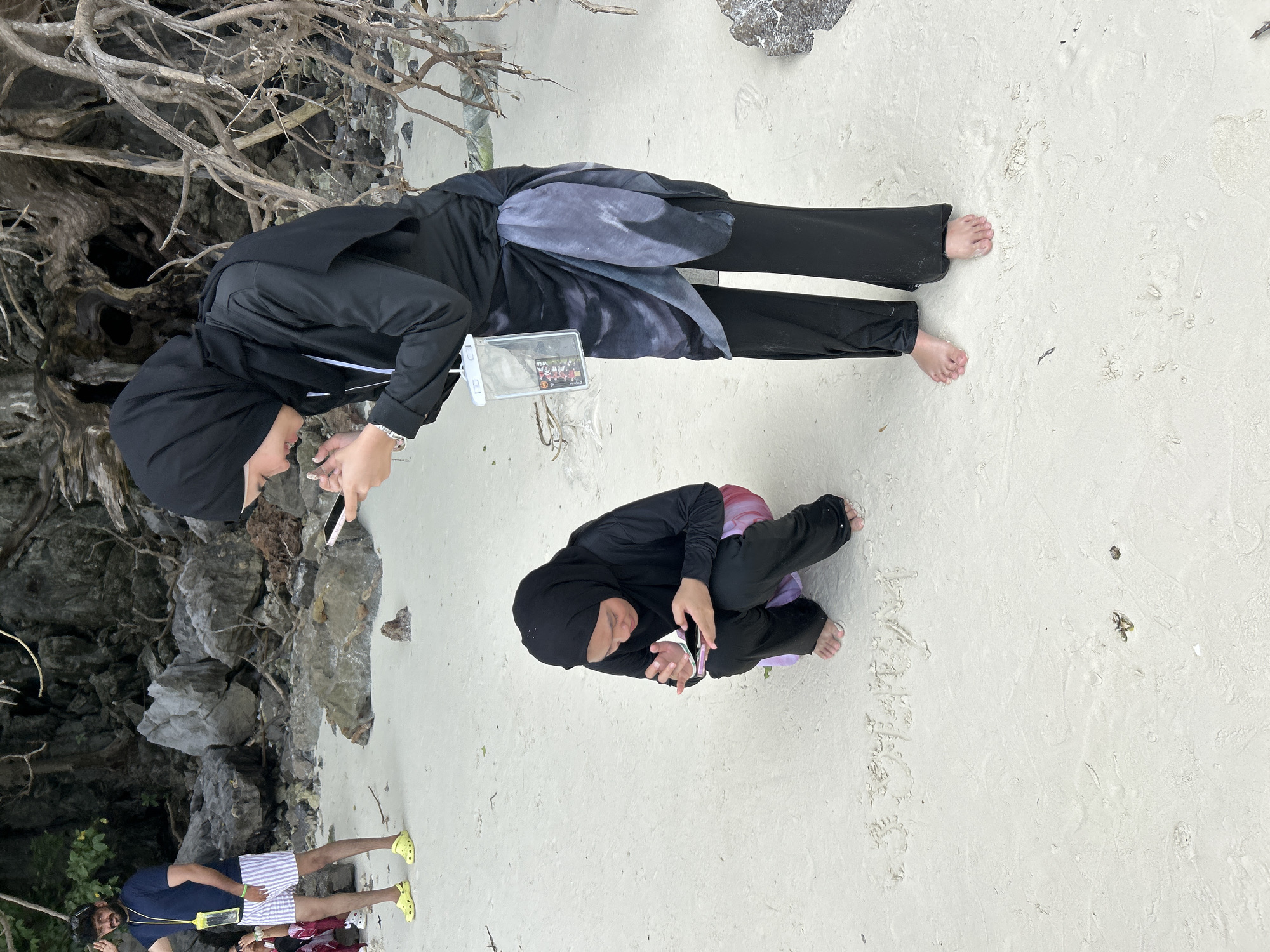 Two individuals are on a sandy beach, likely in Koh Phi Phi, Thailand, based on the characteristic limestone karsts and turquoise waters often found there, though the water is not visible in this frame. The person in the foreground, wearing a black hijab and black modest swimwear, is crouching and intently looking at their phone. They have a pink phone case and appear to be writing in the sand with their finger. Behind them and slightly to the left, a second person, also in a black hijab and modest black swimwear, stands and is looking down at their phone. This person is wearing a blue and white striped sarong tied around their waist and has a clear waterproof phone pouch hanging from their neck. In the background, a third person is visible to the far left, wearing a blue t-shirt and striped shorts, and yellow Crocs. The beach is composed of fine white sand, with scattered rocks and exposed tree roots forming a natural backdrop. The overall scene depicts a casual beach outing with a focus on mobile device usage.