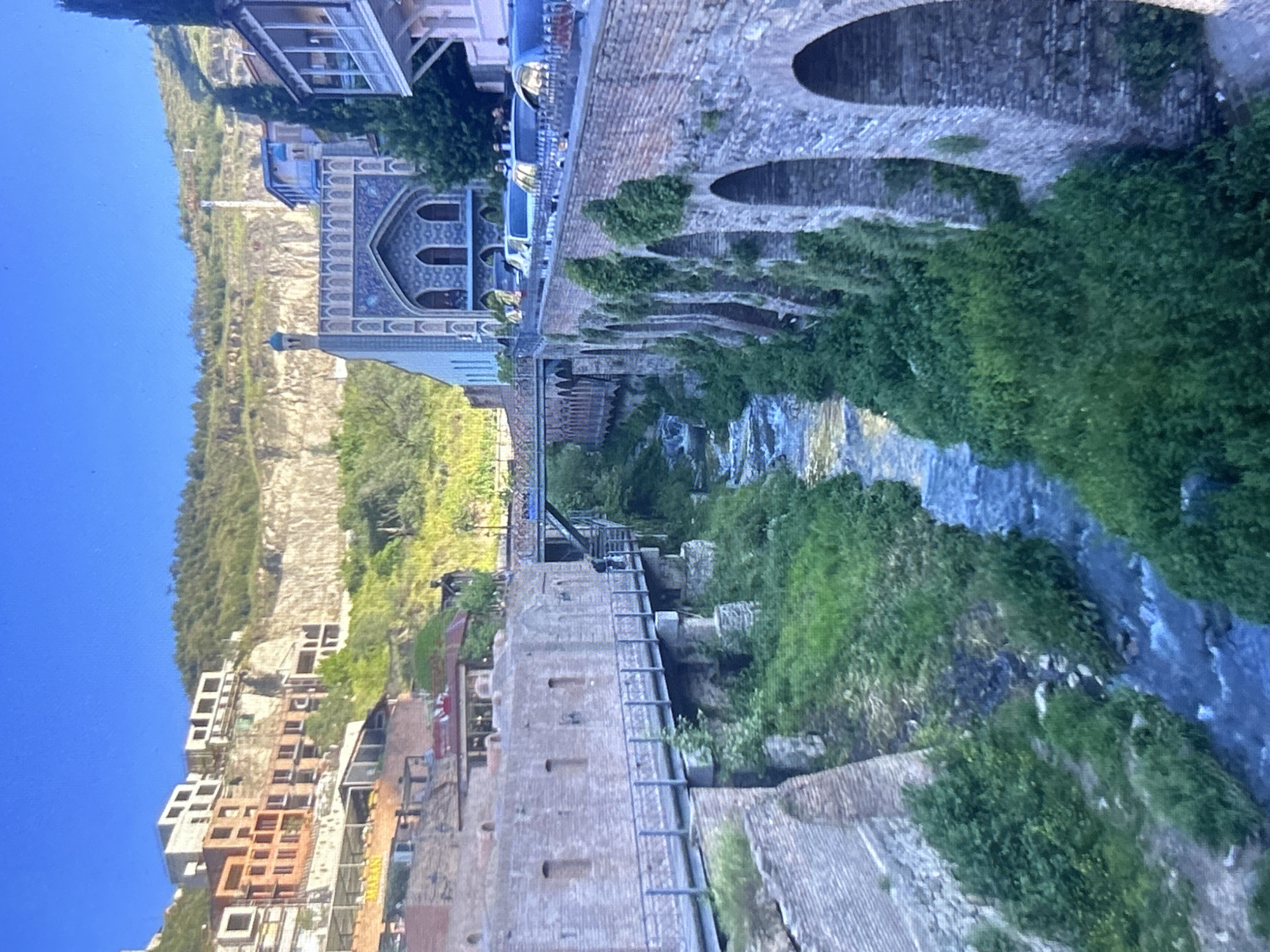 The image depicts a view in Tbilisi, Georgia, showcasing the Leghvtakhevi waterfall and the historic sulfur bath district. A river flows through a narrow gorge, flanked by lush greenery. On the left side of the river, a brick structure with arches is partially covered in vegetation. Across the river, there is a blend of historical and modern buildings, including Orbeliani Baths, with its distinctive blue-tiled facade. The skyline features a steep cliff face with buildings clinging to its slope. The sky is clear and blue.