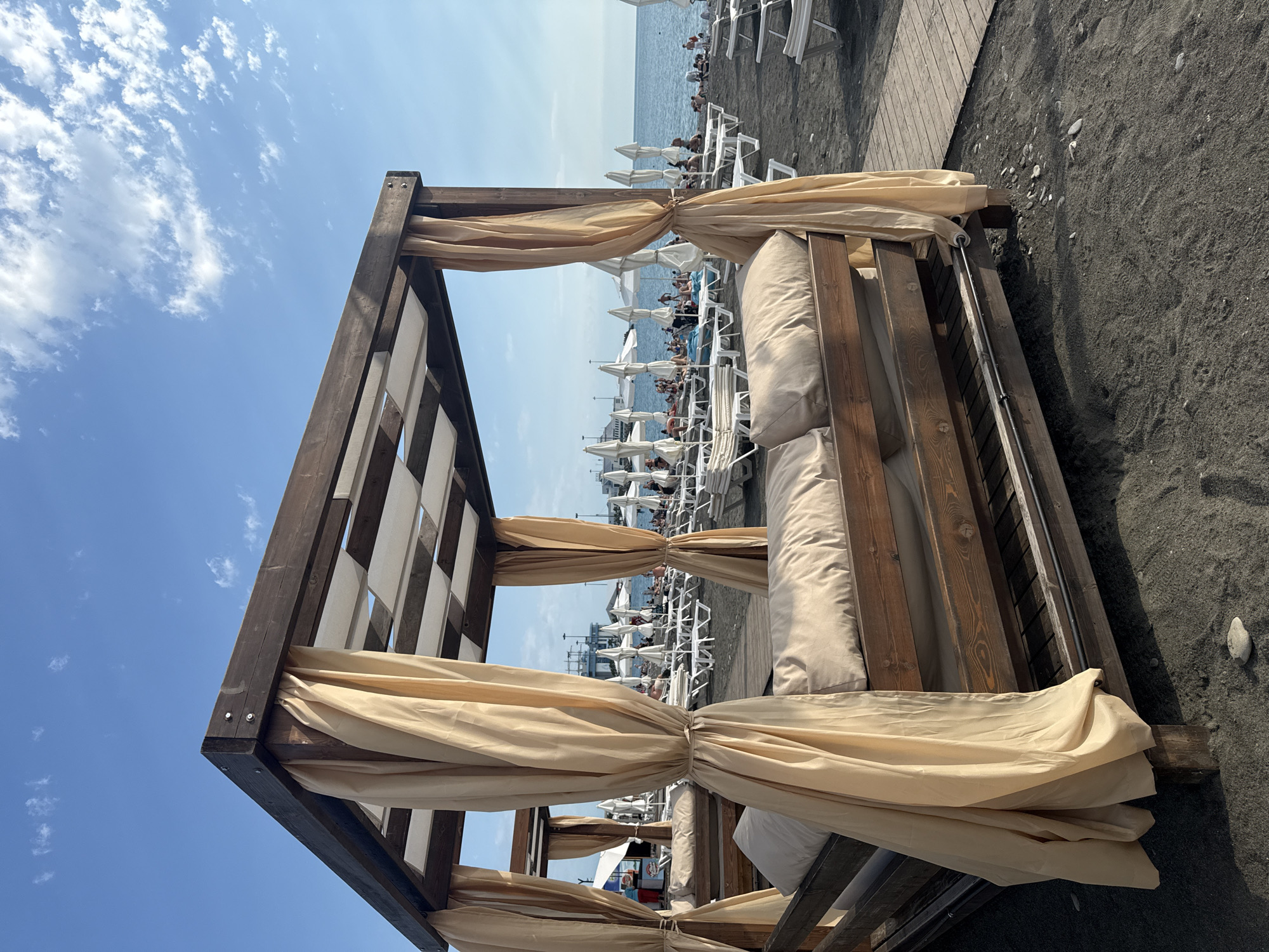 This media file depicts a beach scene in Sochi, Russia. The primary subject is a wooden cabana with beige curtains, providing shade on a sandy beach. Several white lounge chairs are visible behind the cabana, some occupied by people. The sea is in the background, with a clear blue sky overhead dotted with a few wispy clouds. The sun appears to be high in the sky, suggesting it is daytime, likely mid-morning or early afternoon. The mood is relaxed and leisurely, consistent with a beach vacation. The sand is dark, and small pebbles are scattered throughout. There is a wooden walkway leading away from the cabanas, and further back, more lounge chairs and umbrellas are arranged. The overall impression is one of a typical resort beach on a sunny day. No visible text is present in the image.