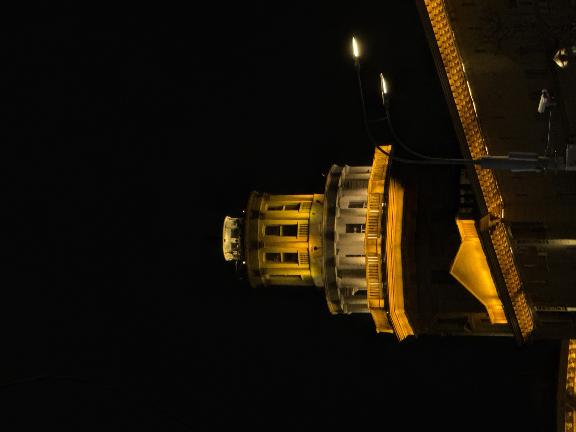 The image depicts a structure illuminated at night. The structure appears to be a tower or part of a bridge, featuring a cylindrical section with windows and decorative elements. Additional structures are present above and to the right, adorned with golden lighting. The backdrop is a dark night sky. Given that the bridge is illuminated and Moscow is known for its well-lit landmarks, the image likely shows one of Moscow's architectural landmarks at night.