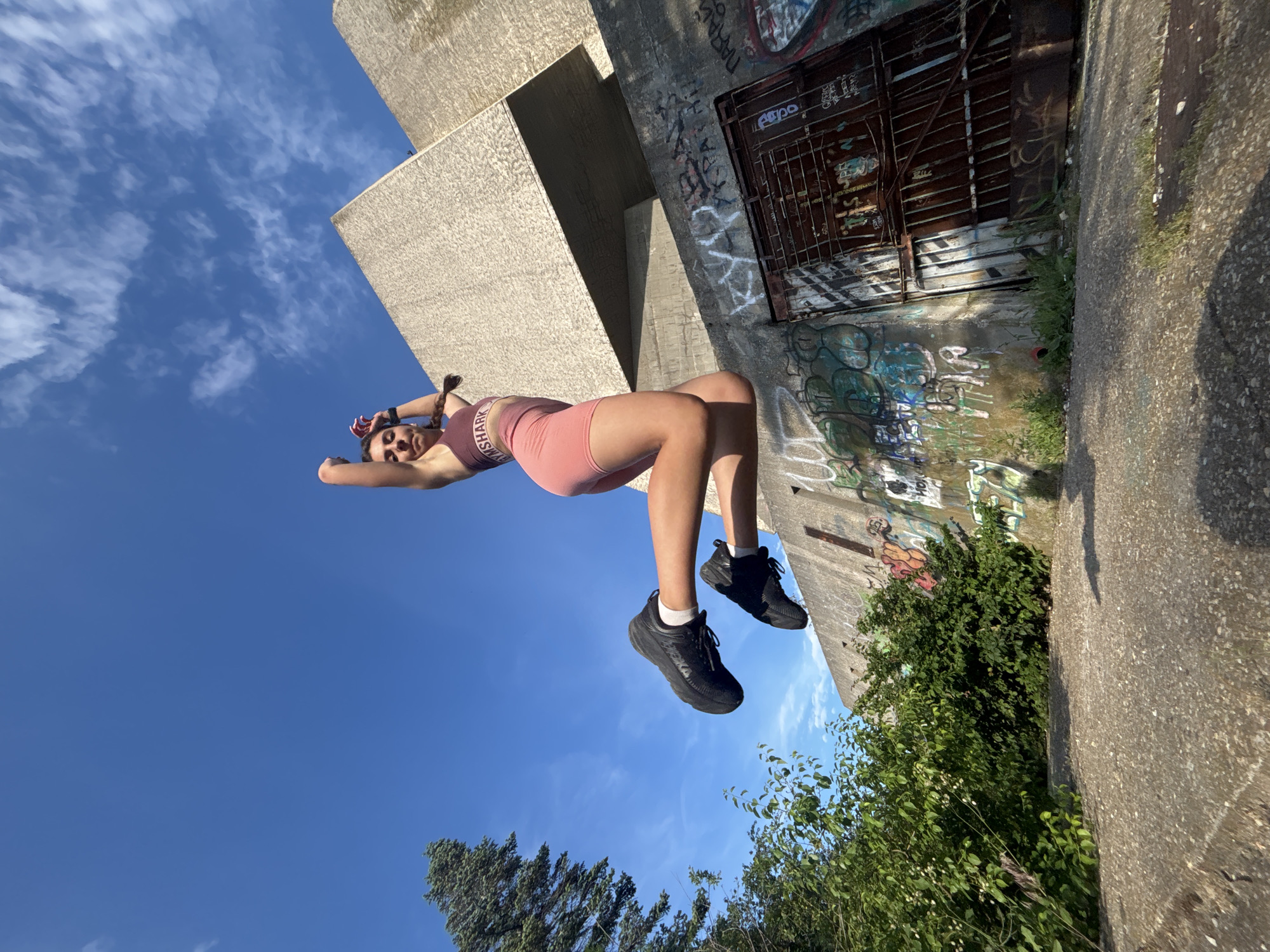 A young woman with brown braided hair is captured mid-air in a powerful jump, her body angled towards the sky. She is wearing a pink sports bra and matching shorts, with black athletic shoes and white socks. Her arms are raised above her head, and her legs are bent, giving the impression of explosive movement.
The background is a striking juxtaposition of natural and man-made elements. Behind her, a large, Brutalist-style concrete structure dominates the upper right portion of the frame. Its rough, textured surface and sharp angles suggest a stark, utilitarian design. Below this structure, a concrete wall is heavily adorned with colorful graffiti, its layers of spray paint creating a chaotic yet vibrant tapestry. To the left of the woman, lush green foliage, including a tall evergreen tree, provides a natural contrast to the urban decay. The ground in the foreground is rough, grey concrete, cracked and uneven, with patches of grass pushing through. The sky is a brilliant, clear blue with a few wispy white clouds, indicating a bright, sunny day.
The overall impression is one of dynamic energy and youthful exuberance set against a backdrop of urban grit and natural resilience. While the specific location within Varna, Bulgaria, is not explicitly indicated by visual cues, the architecture and graffiti suggest an industrial or post-industrial area, possibly on the outskirts of the city.