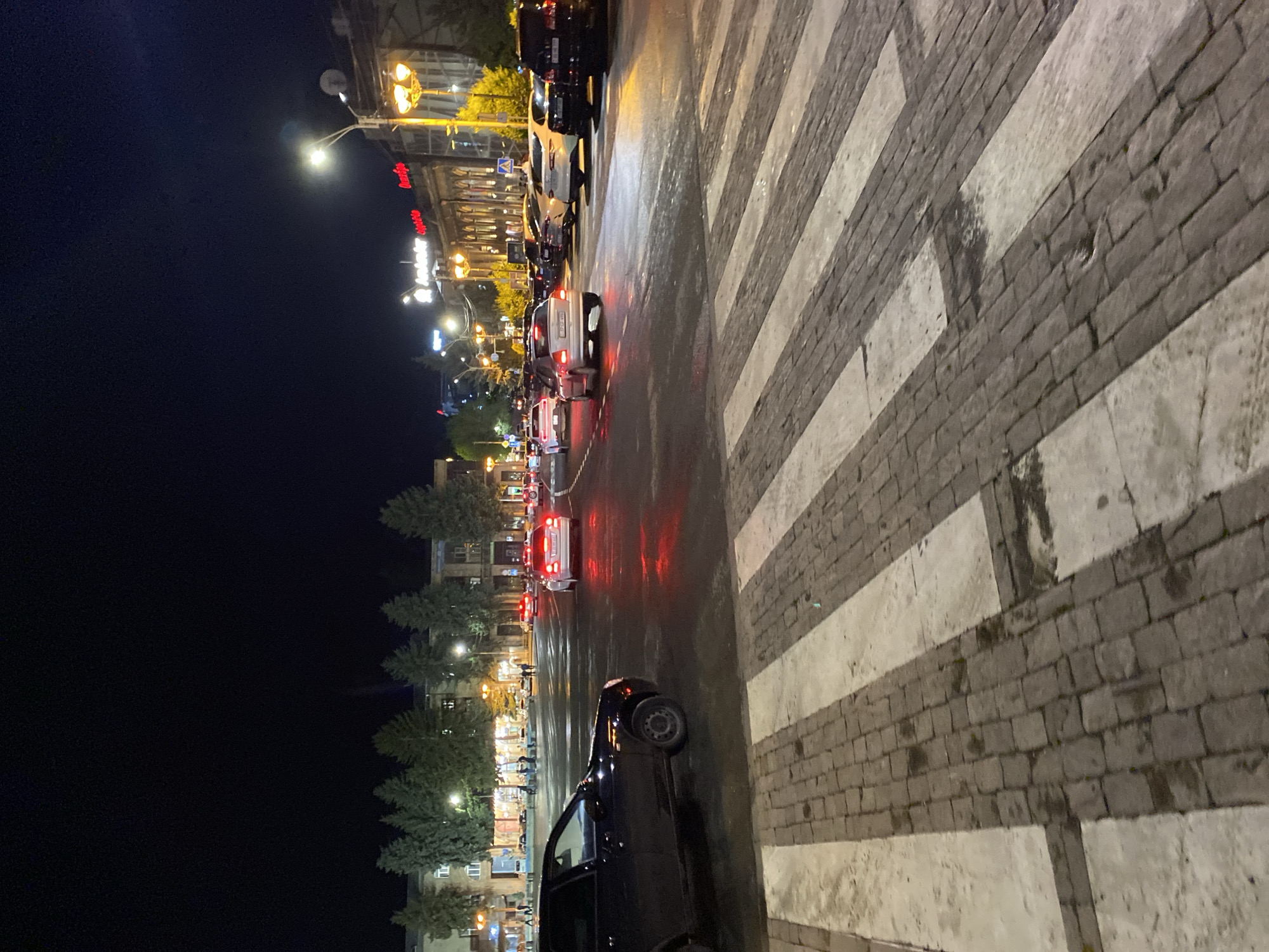 The image depicts a street scene at night in Gyumri, Armenia, likely in the central area given the presence of illuminated buildings and traffic. A pedestrian crossing is prominent in the foreground, paved with cobblestones and white stripes. Several cars are on the wet asphalt road, some with their brake lights illuminated, indicating slow movement or stopping. On the left side, a dark colored car is parked at an angle. In the background, buildings line the street, featuring illuminated windows and storefronts, with trees in between some of them. Streetlights cast a warm yellow glow on the scene, reflecting on the wet road surface. The sky is dark, characteristic of nighttime. There are no clear individuals identifiable in the foreground, though blurry figures can be seen in the mid-ground near the buildings. The overall impression is a moderately busy urban street at night.