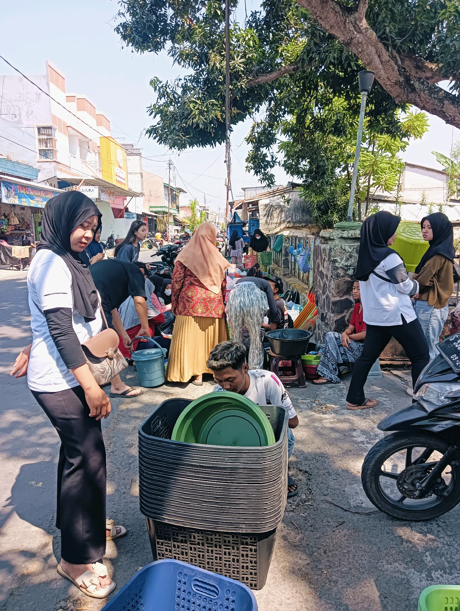 This is a street scene in Ploso, Indonesia, on a sunny day. The foreground is dominated by a stack of black plastic baskets, some containing green plastic basins. A person wearing a white long-sleeved shirt and black pants is standing to the left, looking down. To their right, a person wearing a red and gold patterned top and a tan hijab is standing. Further to the right, two young women in hijabs are conversing. In the background, there are buildings, trees, and a parked motorcycle. People are engaged in various activities, suggesting a market or busy street environment. The ground is paved asphalt.