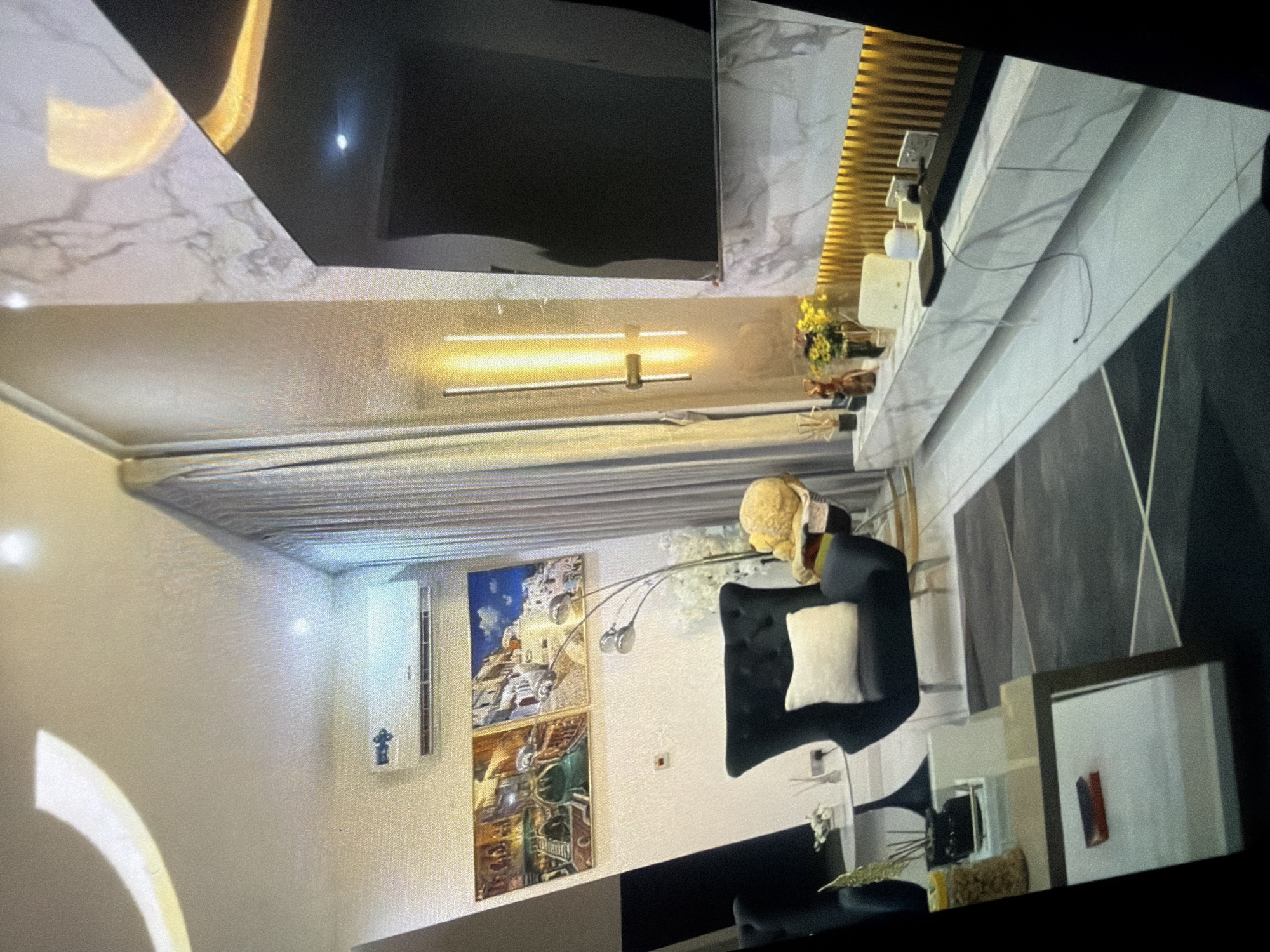 The image depicts a modern living room with a focus on a large television mounted on a marble-clad wall. A minimalist entertainment unit with a white marble finish runs along the base of the wall, adorned with decorative items and electronics. To the left of the television, a contemporary floor lamp with multiple arms is positioned near a dark velvet armchair. Above the armchair, two framed artworks are hung on the wall, depicting architectural scenes. An air conditioning unit is visible on the wall above the artwork. A contemporary coffee table with a reflective surface is placed on a geometric-patterned rug in the foreground, with a decorative jar of snacks on its surface. The overall aesthetic is sleek and sophisticated, with a blend of modern furniture and decorative elements. The scene is illuminated by a vertical wall sconce, the television screen, and ambient ceiling lighting, creating a warm and inviting atmosphere.