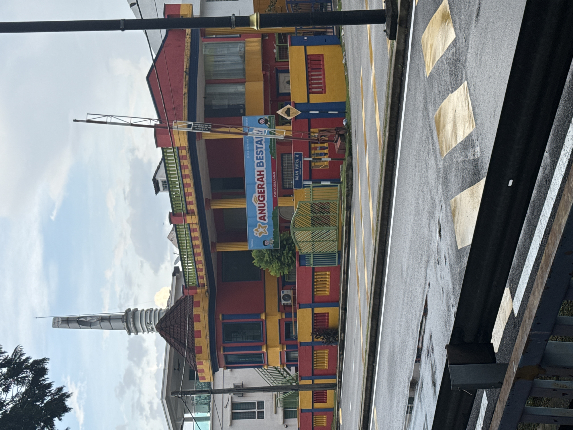 The image captures a scene in Ampang Jaya Municipal Council, Malaysia. A brightly colored building, predominantly red, yellow, and blue, stands adjacent to a road. A sign reads "ANUGERAH BESTA" in blue lettering. There's also a street sign indicating "JALAN PUTRA 4" and another sign indicating that the MADKA school is in the vicinity. In the background is a tall, white minaret and there is a small tree to the left of the minaret. The road appears wet, marked with yellow lines and a series of diamond-shaped markings. Part of a grey metal structure, potentially a bridge or railing, is visible in the foreground.