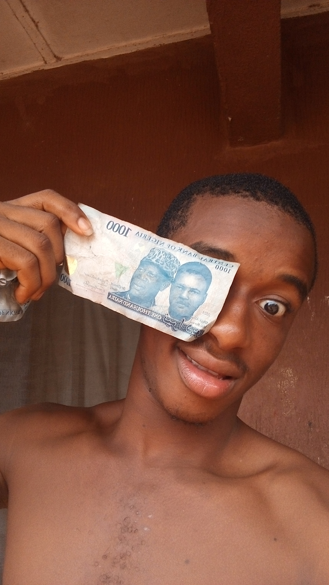 The image features a close-up portrait of a young man, likely in Onitsha, Nigeria, given the visible currency and provided context. He has dark skin and short dark hair. The man is shirtless, suggesting an informal, indoor setting. He holds a crumpled Nigerian 1000 Naira banknote over his left eye. His right eye is wide open, and he displays a playful grin or smile on his face, conveying a cheerful and possibly humorous mood.
The background consists of a plain reddish-brown wall or interior surface, with subtle lines and textures indicating an architectural element in the upper corners. The lighting appears soft and diffused, characteristic of indoor natural light.
The Nigerian 1000 Naira banknote is a prominent object. It is slightly creased and displays the denomination "1000" in numerals, along with the text "CENTRAL BANK OF NIGERIA" and "ONE THOUSAND NAIRA." Arabic script is also visible on the note. The portraits of Alhaji Aliyu Mai-Bornu and Dr. Clement Isong, former governors of the Central Bank of Nigeria, are clearly depicted on the banknote. The man's hand is visible on the left, holding the currency.