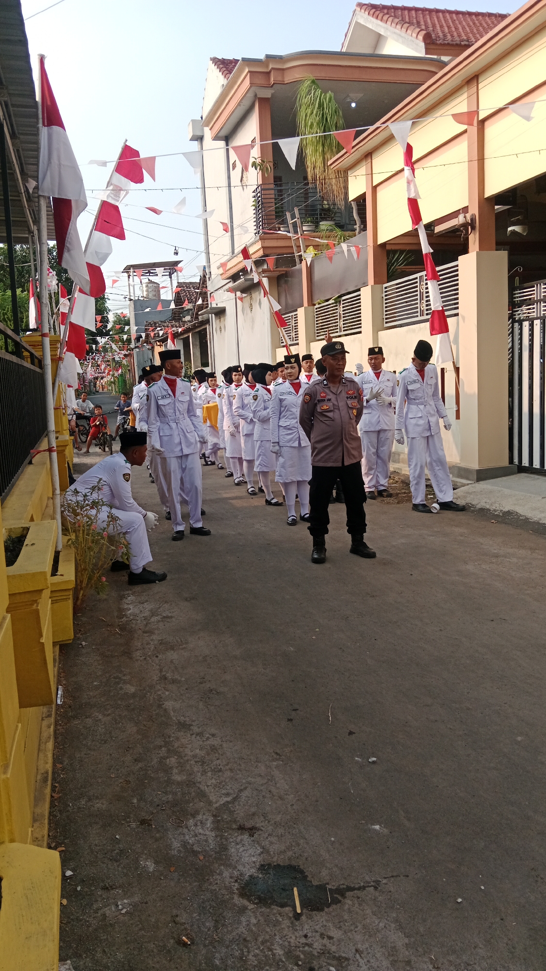 A police officer stands in the center of a paved street in Paiton, Indonesia, facing forward with hands clasped behind his back. He is wearing a grey uniform with a badge on his chest. To his right, a group of young people in white uniforms stand in a line, facing the same direction as the officer. To the officer's left, another group of young people in white uniforms are also lined up. One person in a white uniform is kneeling on the side of the street. The street is decorated with red and white flags and pennants. Buildings line the street, some with balconies. The sky is clear and bright.