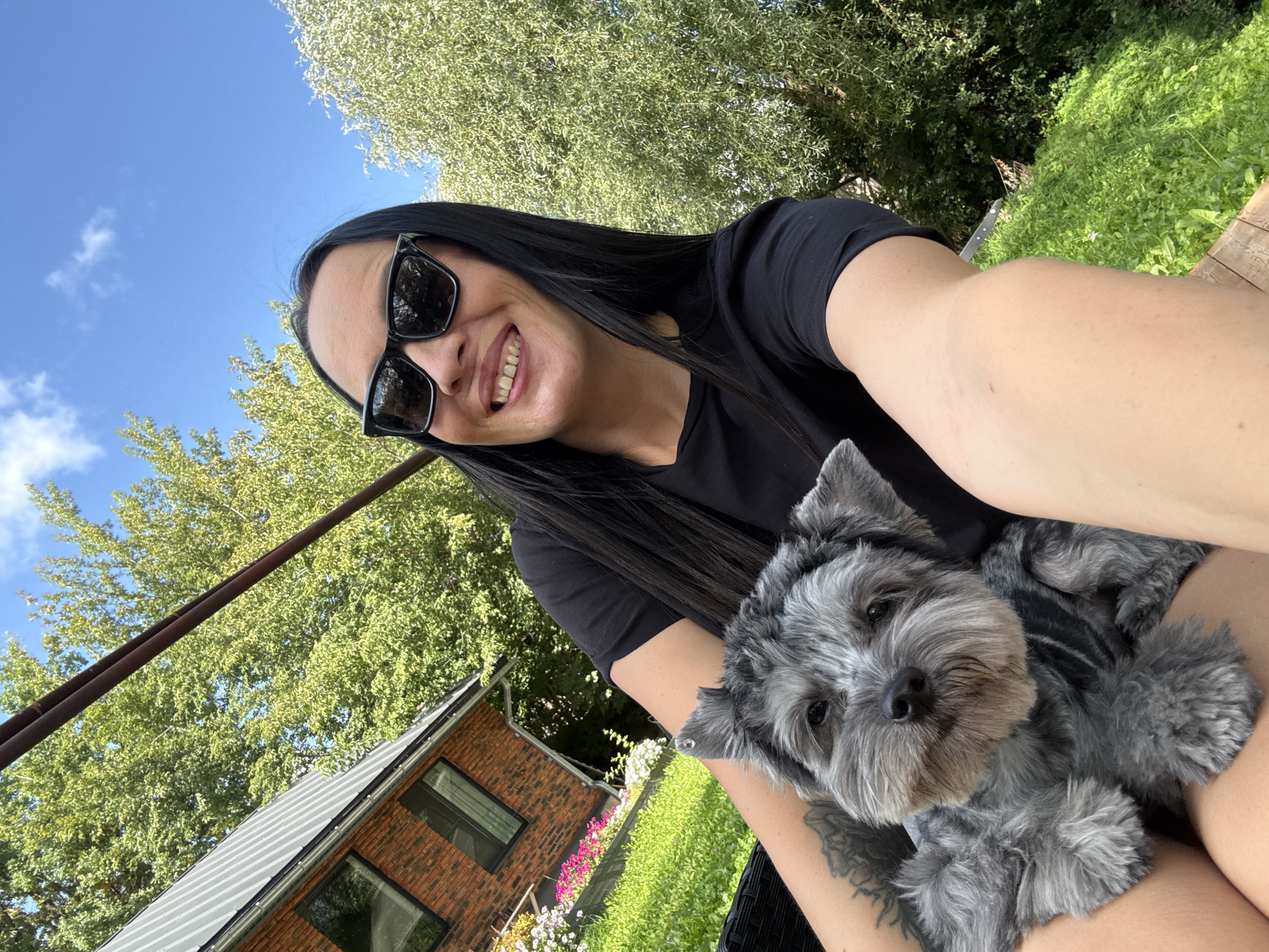 A woman with long black hair and sunglasses smiles for a selfie with a small grey dog in her arms. They are outdoors, with lush green trees and a bright blue sky with a few white clouds visible in the background. To the left, part of a red brick house with a grey roof and dark windows is visible. There are also colorful flowers planted in front of the house. The scene appears to be a residential area on a sunny day.