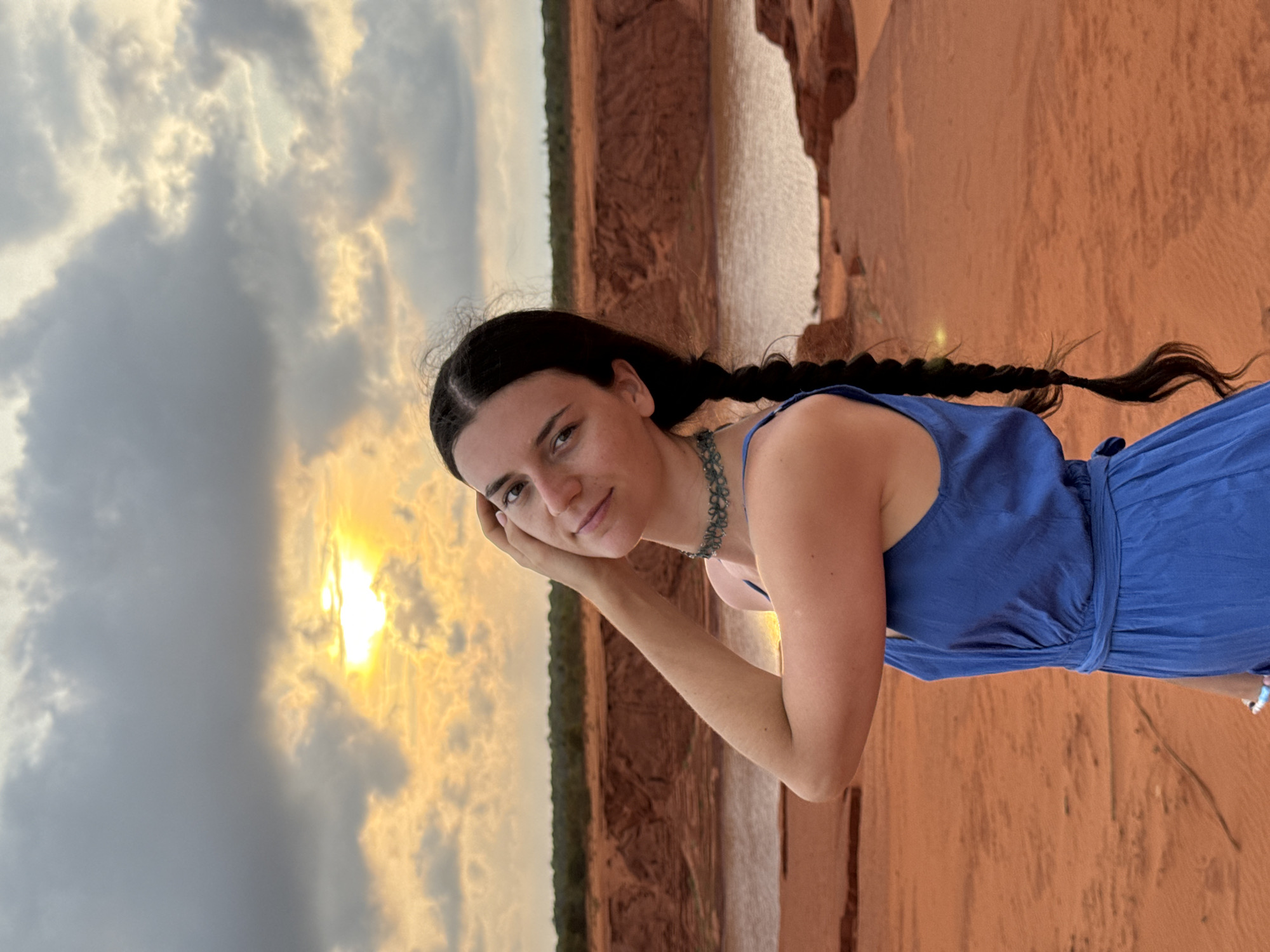 A woman with dark hair styled in two braids stands in front of a landscape at sunset. She is wearing a blue dress and a necklace. The background features a body of water bordered by red cliffs and foliage, typical of the geological formations in Mui Ne, Vietnam. The sky is filled with clouds and the setting sun creates a golden glow.
