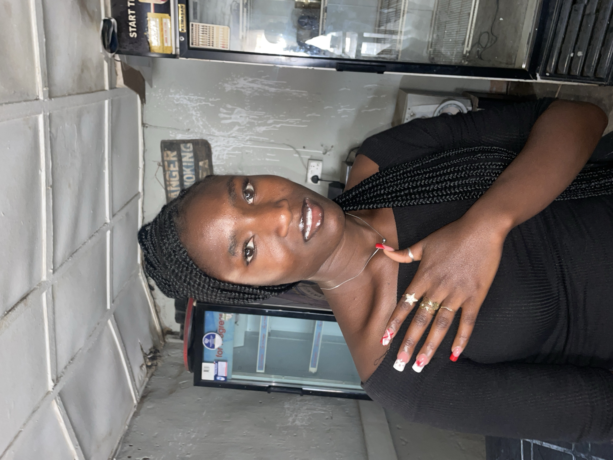 A portrait-style photograph features a young Black woman looking directly at the camera with a slight smile, set in what appears to be an indoor commercial space in Jos, Nigeria. She has dark skin, braided hair pulled back, and wears a black off-the-shoulder garment. Her lips are glossy, and she sports a silver chain necklace. Her right hand, resting near her chest, displays an elaborate manicure with red and white French tips, some adorned with small hearts or stars, along with several rings including a star-shaped ring and plain gold bands.
The background suggests a shop or bar environment. To her left, there's a light-colored, textured wall, possibly painted cinder blocks or large tiles, showing some wear. Behind her, multiple glass-fronted commercial refrigerators or display cases are visible, indicative of a retail setting. One refrigerator bears the brand name "KLIMASAN" and a label with "for gress" and a blue circular logo. Above and to her right, a black sign partially reads "START T", and a weathered sign further back clearly states "DANGER SMOKING". An electrical outlet is also visible on the wall. The lighting appears to be artificial, casting a casual ambiance over the scene.