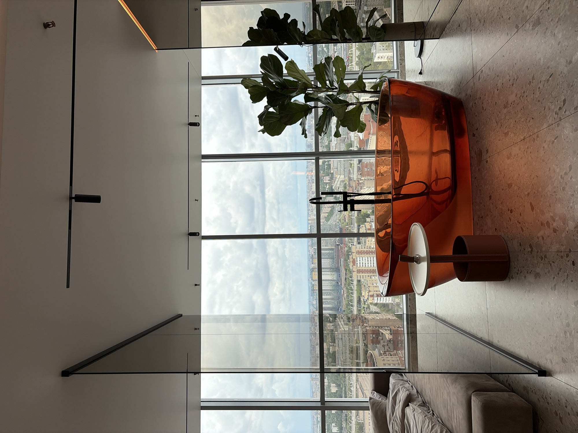 This is a medium shot, eye-level, interior photograph of a bedroom with an en-suite bathroom area, overlooking a city skyline. The room is modern and luxurious, with large floor-to-ceiling windows providing a panoramic view of Saint Petersburg, Russia. The sky is overcast with large, fluffy clouds.
Dominating the center of the image is a vibrant orange, free-standing bathtub. It has a sleek, contemporary design and appears to be made of a translucent material. A matte black faucet with a single handle is installed in the tub. To the left of the bathtub is a small, round side table with a white top and a brown leather cylindrical base.
To the left of the bathtub, partially visible, is a large bed with light-colored linens. A floor-to-ceiling glass partition separates the bedroom area from the bathroom area. To the right of the bathtub stands a tall, leafy green plant in a planter. The floor is a speckled terrazzo material. The ceiling features a track lighting system. The overall impression is one of sophisticated urban living.