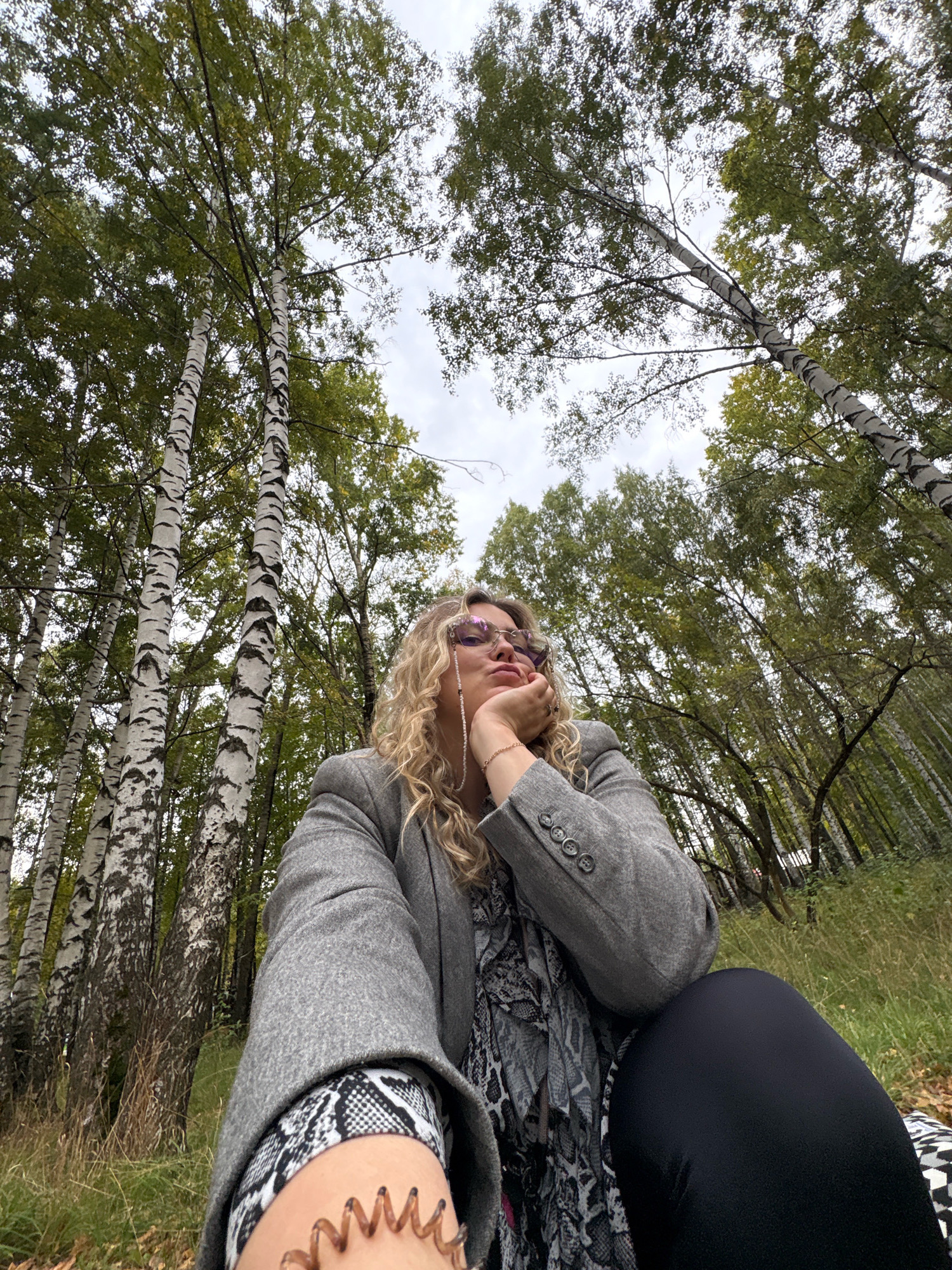 An outdoor, low-angle, medium shot captures a person sitting in a park-like setting with birch trees overhead. The person, facing left, has blonde curly hair and is wearing a gray blazer over a patterned scarf and black leggings. They are resting their chin on their hand and looking slightly upwards. The background consists of a dense forest of birch trees and other foliage, with a cloudy sky visible through the canopy. The ground is covered in dry grass. The image is set in the city of Chaikovsky, Russia, as indicated by the presence of birch trees which are common in the region.
