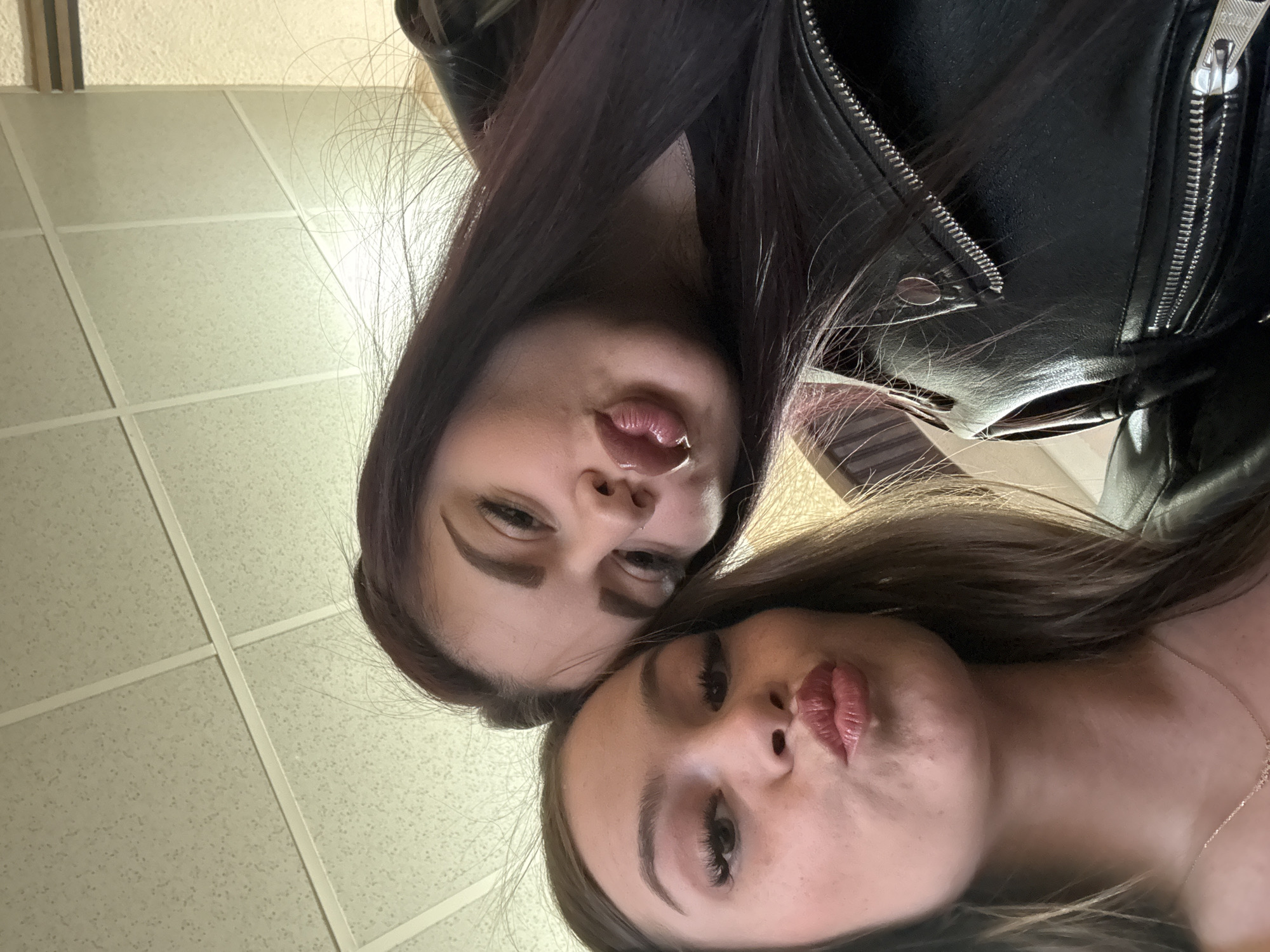 Two young women are posing for a selfie. The woman at the top of the frame has dark hair and a nose ring. She is wearing a black leather jacket. The woman at the bottom of the frame has brown hair and is wearing a necklace. The background appears to be an office building with a drop ceiling. The location in the city of Parvomay, Bulgaria is unknown.