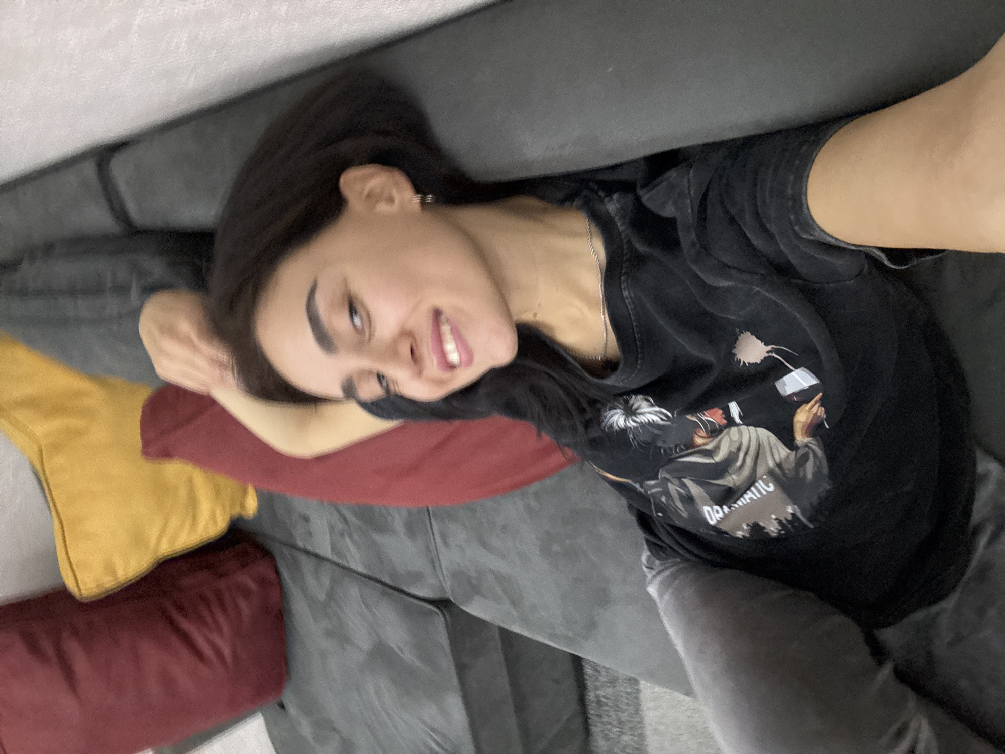 A young woman with dark hair and full lips is lying on a dark gray sofa. She is wearing a black t-shirt with a graphic print on it. There are several pillows on the sofa; one is maroon, and another is yellow. The woman is looking at the camera with a slight smile. The image appears to be taken indoors, in a residential setting, and there are no visual cues to suggest the location is in Tbilisi, Georgia.