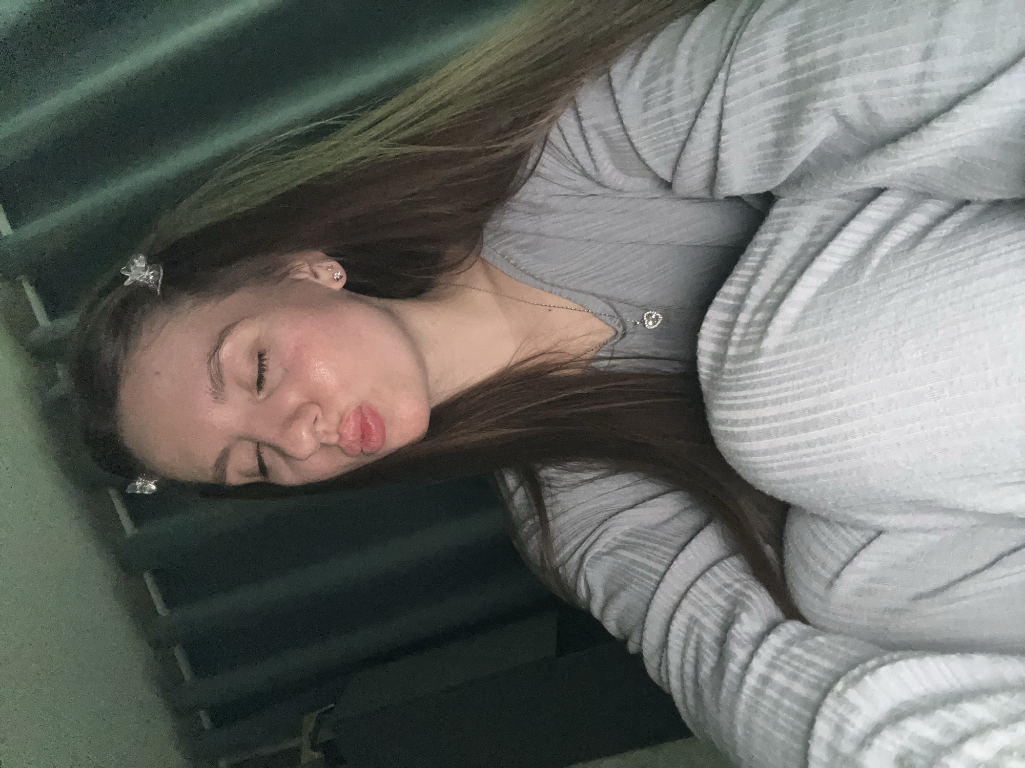 A young woman with long, straight brown hair is depicted in a diagonal, close-up shot, appearing to take a selfie. She is wearing a light gray, ribbed, long-sleeved top, and a similar textured fabric, possibly a blanket or matching bottoms, covers her lower body. Her hair is adorned with two small, clear, butterfly-shaped clips near her temple. She wears small, sparkly stud earrings and a delicate silver necklace with a heart-shaped pendant. Her eyes are closed, and her lips are pursed into a playful "kissing face" expression. The background is dark and indistinct, featuring subtle horizontal lines, suggesting an indoor setting like a bedroom. The lighting is soft, illuminating her face and the textures of her clothing. The image conveys a casual, cozy, and playful mood, presumably taken in Klin, Russia.
