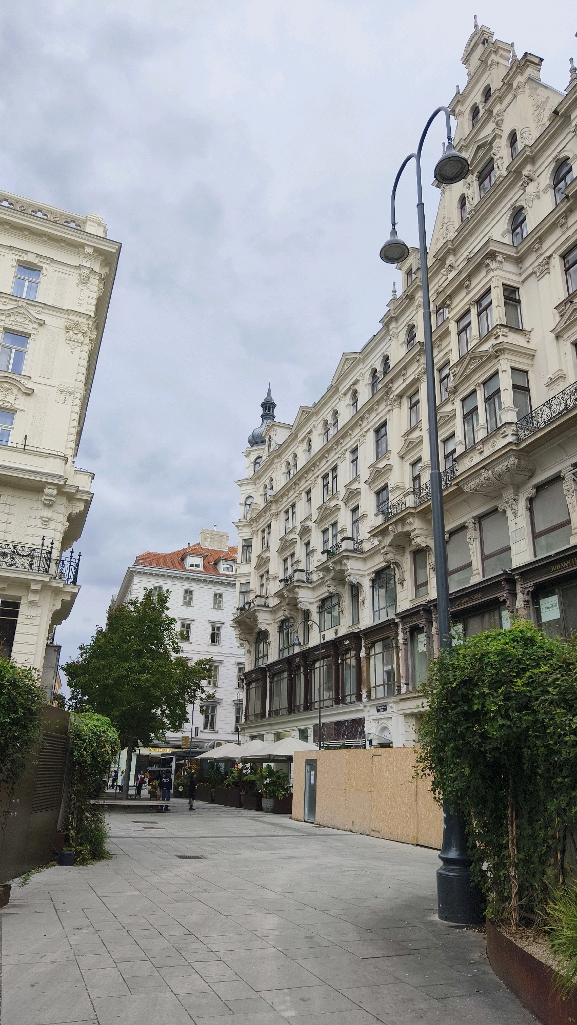 A pedestrian pathway with light gray paving stones leads into a city street lined with ornate, light-colored buildings. A tall, dark lamppost with two curved arms and lamps stands on the right side of the pathway. In the background, to the left, is a building with a mansard roof and white facade, partially obscured by a large green tree with dense foliage. Adjacent to this is another building with a prominent red tiled roof and white walls. On the right, a multi-story building with a cream-colored facade, classical architectural details, bay windows, and wrought-iron balconies dominates the scene. The ground floor of this building features shops with large display windows. Across the pathway, some outdoor seating areas with umbrellas are set up in front of a business. A long, temporary wall constructed of plywood panels runs along the side of the pathway, covering an unknown area. The sky is overcast with diffused light. There are a few people visible in the distance, walking along the pathway and near the outdoor seating area, but their features are not discernible. The overall impression is a scene in an urban European setting, likely a historic district. Based on the architectural style and the presence of establishments like "JOHANN FRANZ," it is highly probable that this location is in Vienna, Austria.